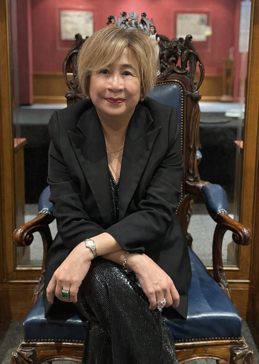 A woman with short blonde hair wearing a white collared shirt and black blazer, looking at the camera with a confident expression, in a modern indoor setting with blurred background lights.