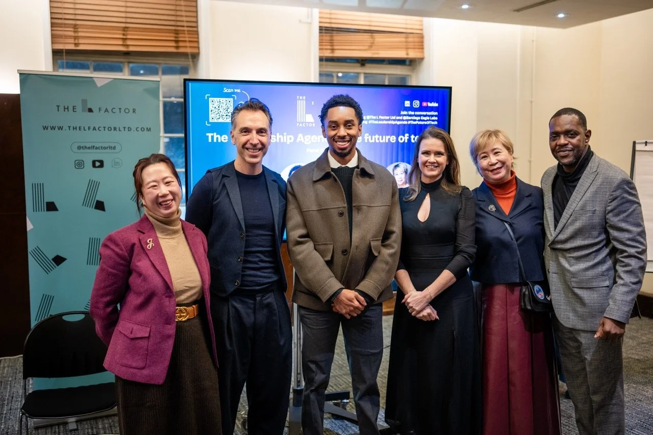 Six people standing together at an event, smiling at the camera with a large screen and a banner in the background.