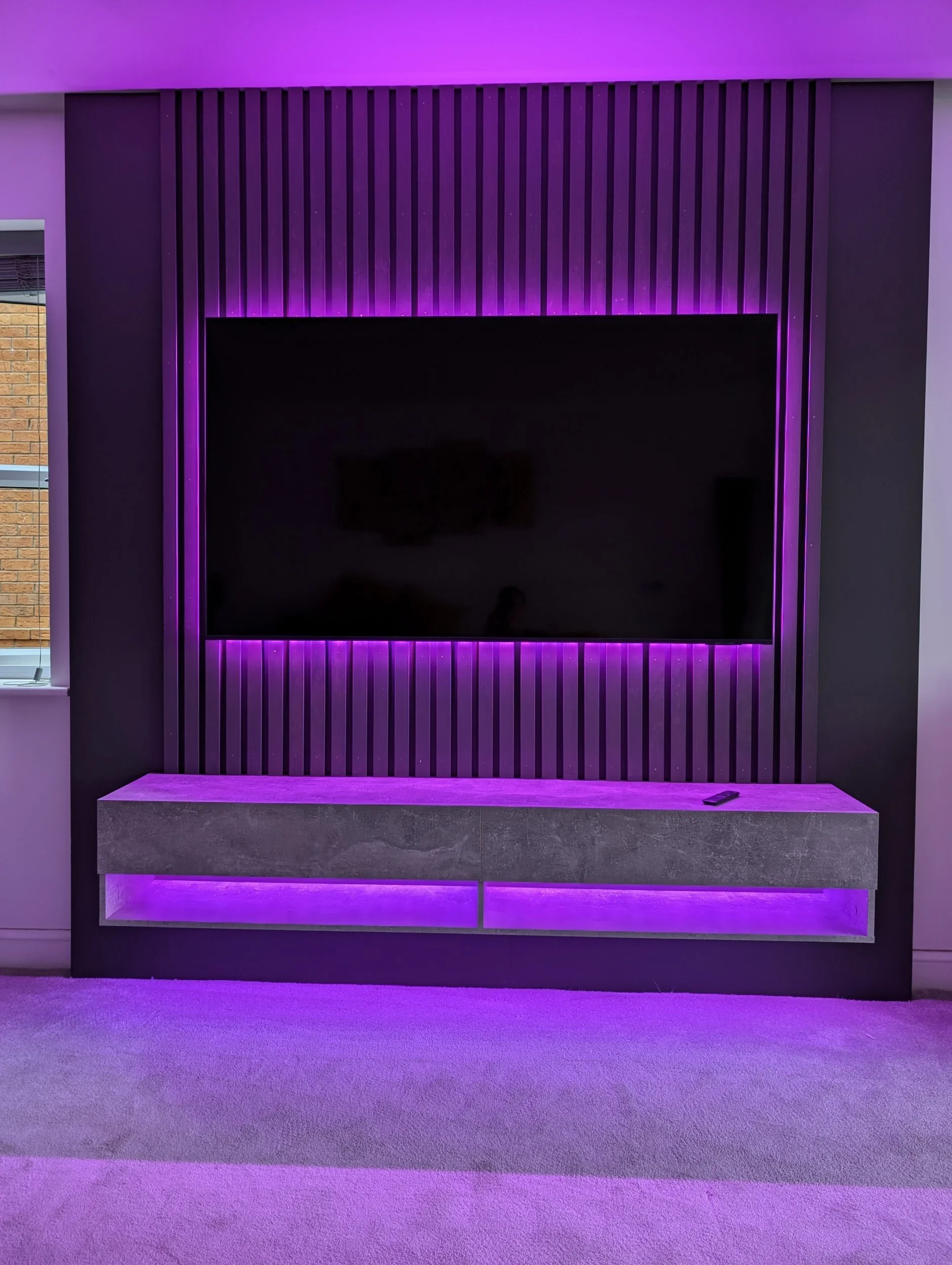 Modern living room entertainment setup with a flat-screen TV mounted on a wall with vertical wooden slats, illuminated by purple LED lighting. A concrete floating media console with purple backlighting and a remote control on top.