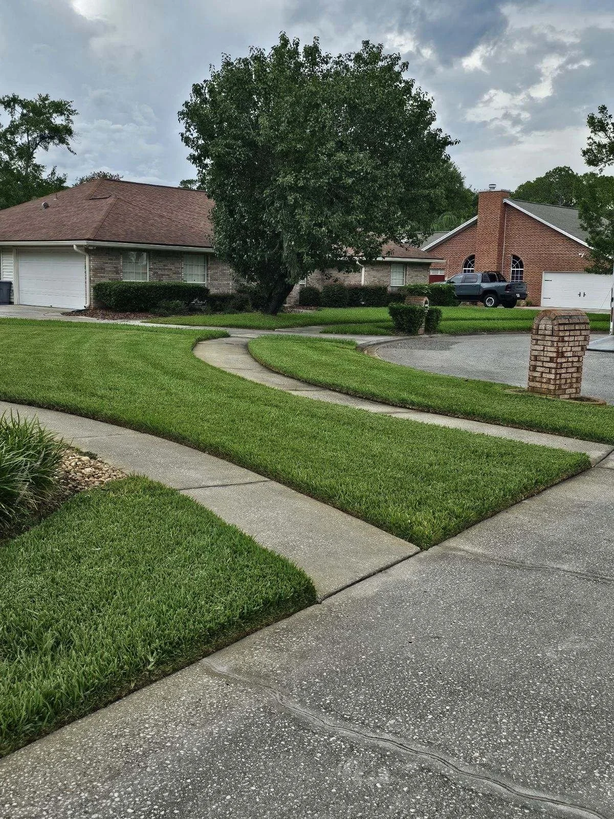 View of a suburban house with a brick and stone exterior, a large tree in the front yard, well-maintained green grass, a concrete sidewalk, and a driveway with a parked pickup truck.