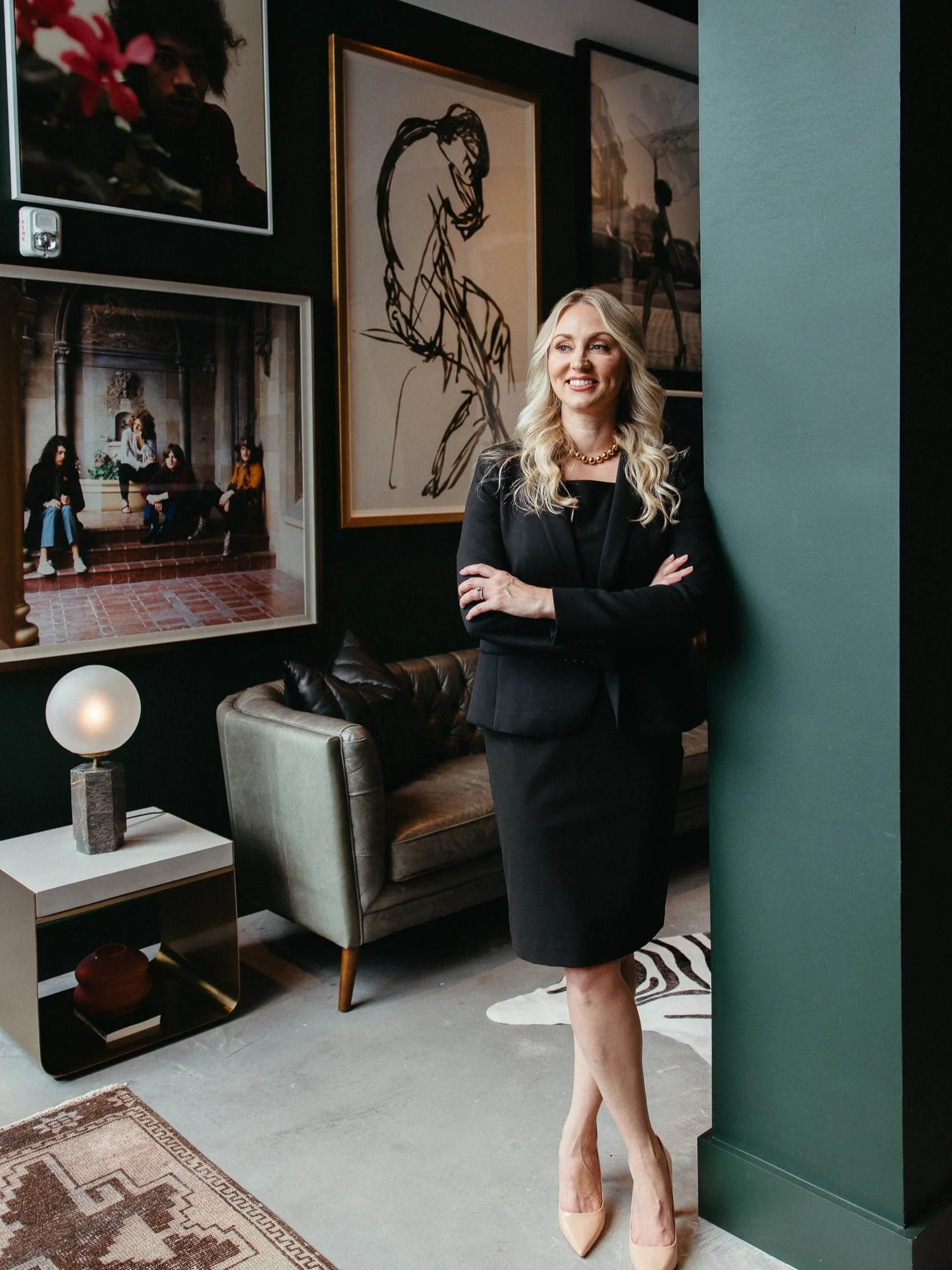 Whitney, the accomplished personal injury lawyer, confidently stands before a wall adorned with pictures in her professional business suit.