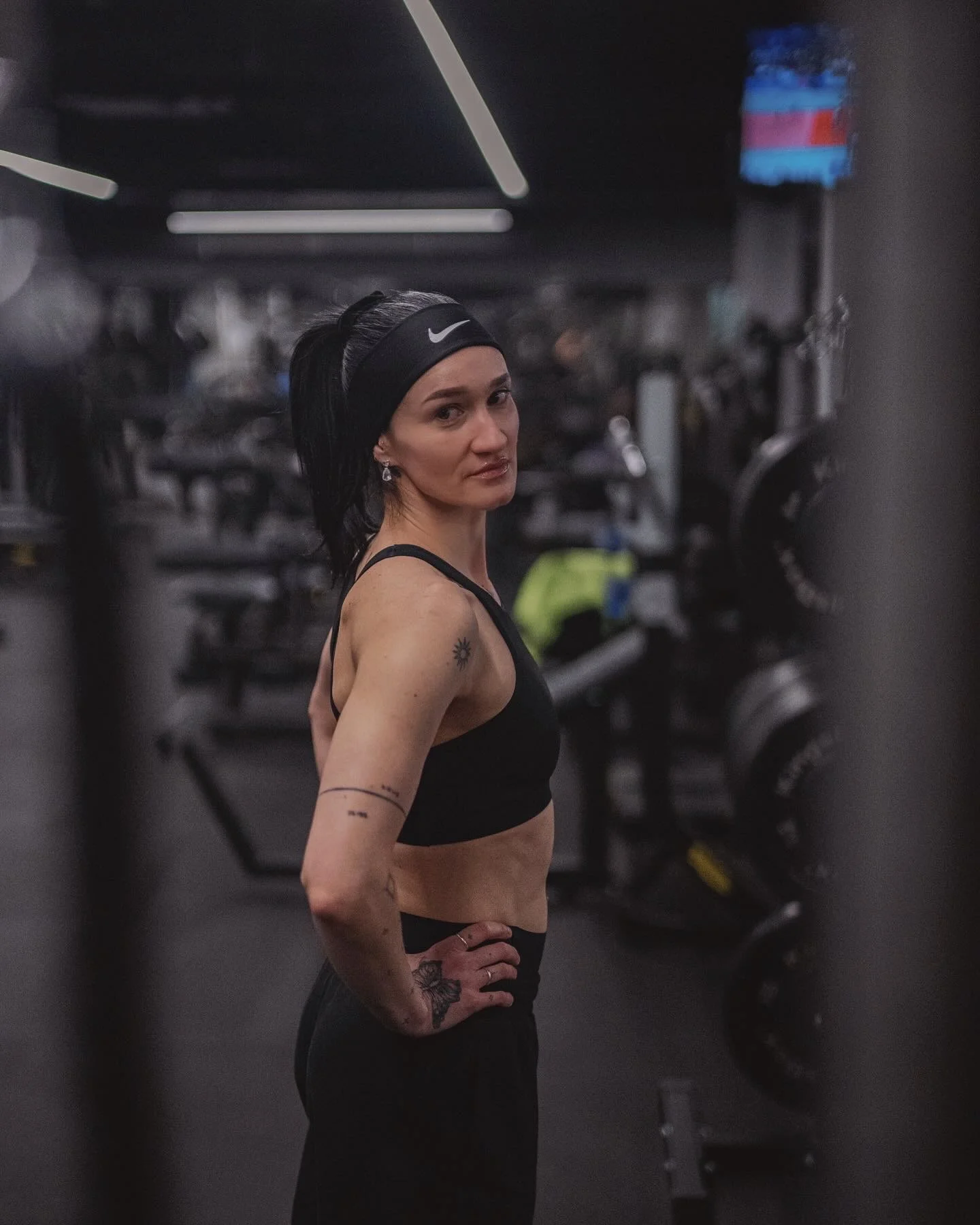 A person in a gym wearing a black sports bra and headband, standing with hands on hips. The background is filled with gym equipment.