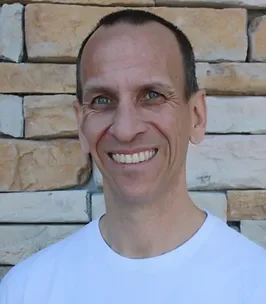 A smiling man with short brown hair and blue eyes, wearing a white t-shirt, standing in front of a stone wall.