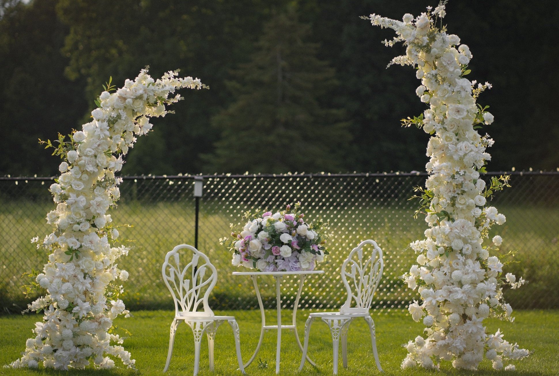 A small outdoor setup with a white metal table and two chairs, adorned with a colorful floral centerpiece. The scene is framed by two large floral arches made of white flowers, set against a background of green grass and a chain-link fence.