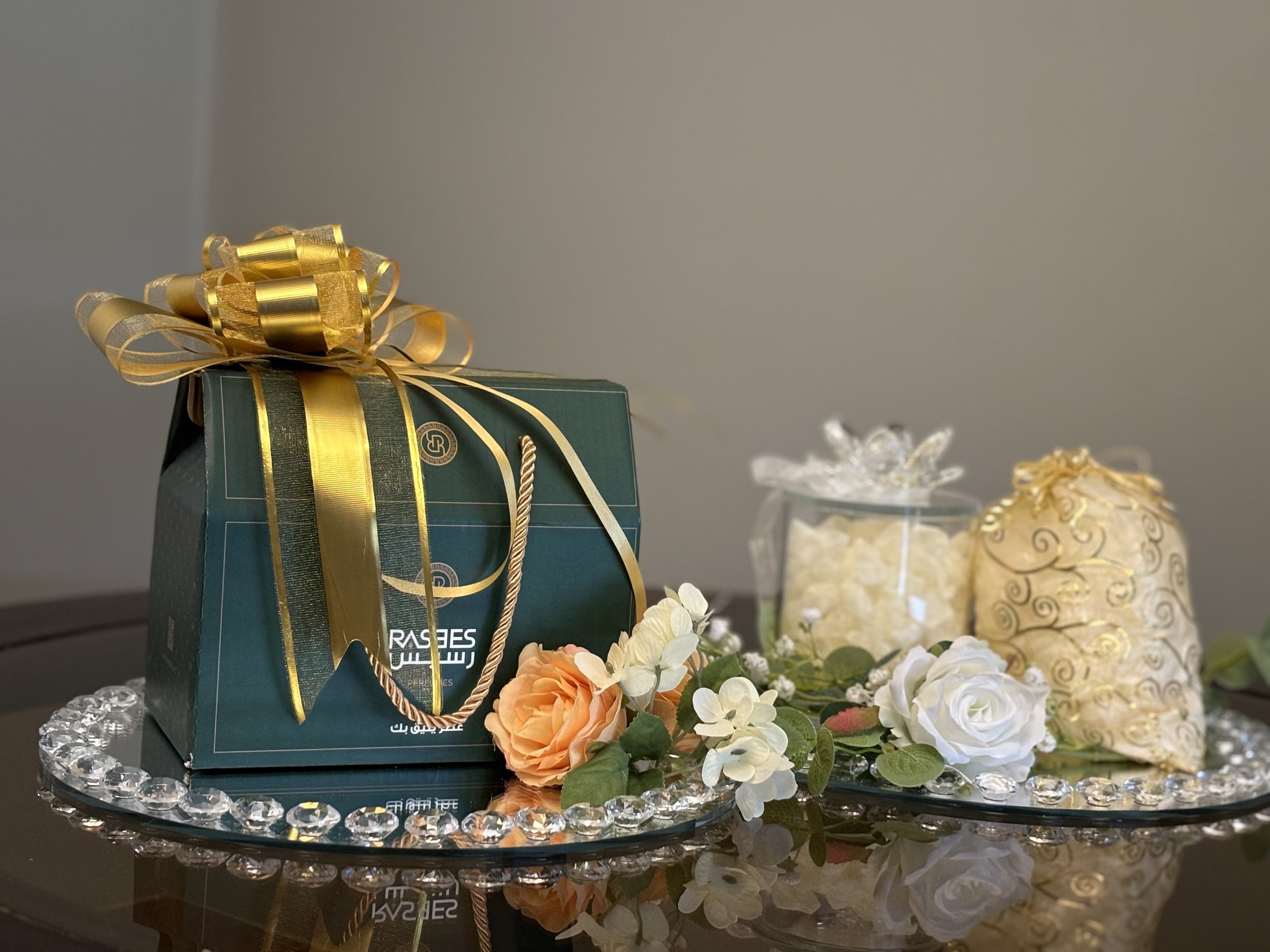 Decorative setup with a green gift box tied with a gold ribbon, surrounded by artificial flowers, a jar of white treats, and a gold-patterned package, all arranged on a glass tray with crystal accents.
