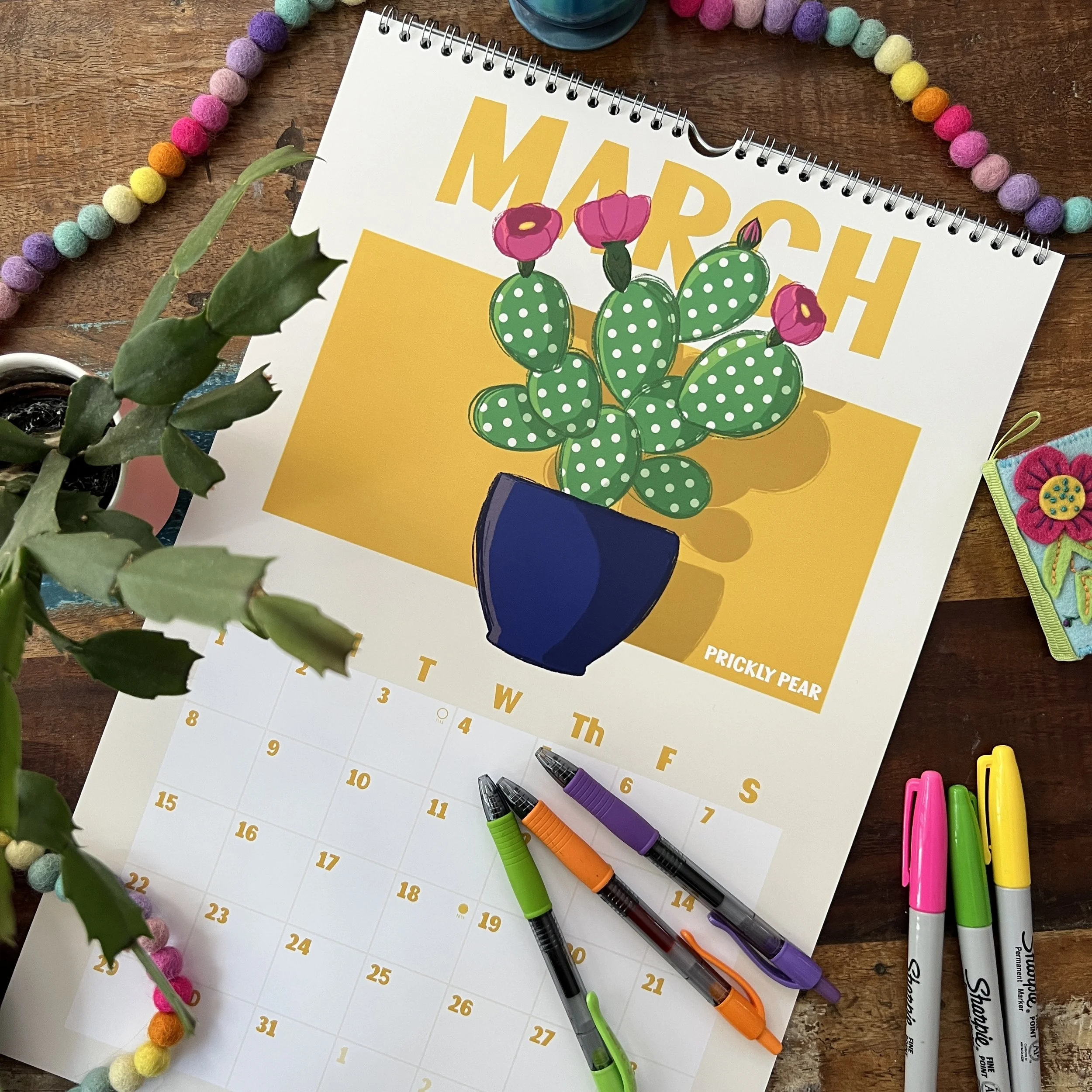 A desk with a colorful paper craft cactus, a calendar page, multicolored markers, a plant, a colorful pom-pom garland, and other craft supplies.