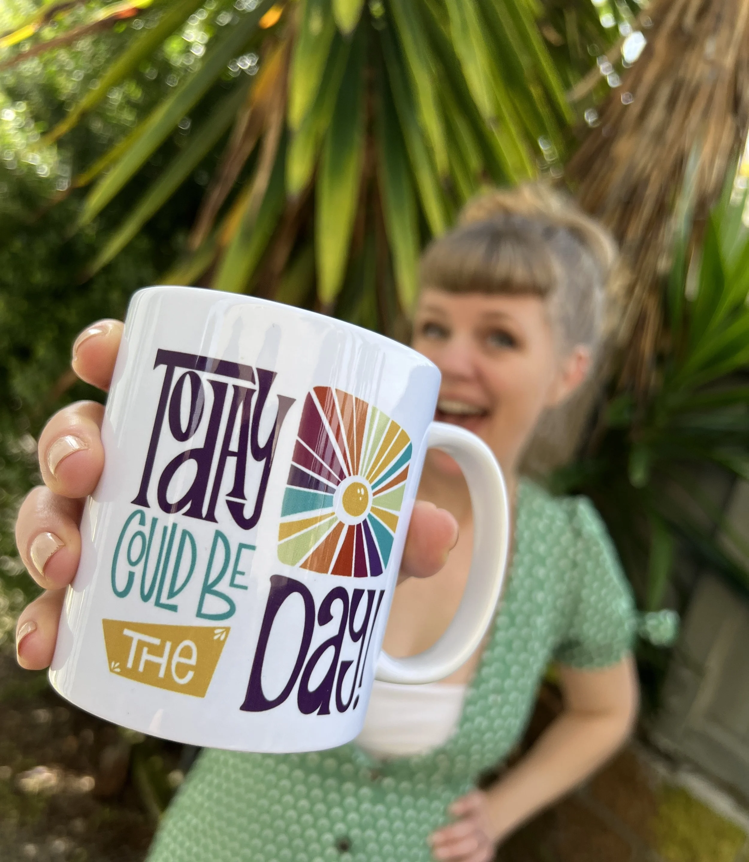 Child with short gray hair holding a white mug with colorful text and a rainbow design, standing outdoors with green plants in the background.