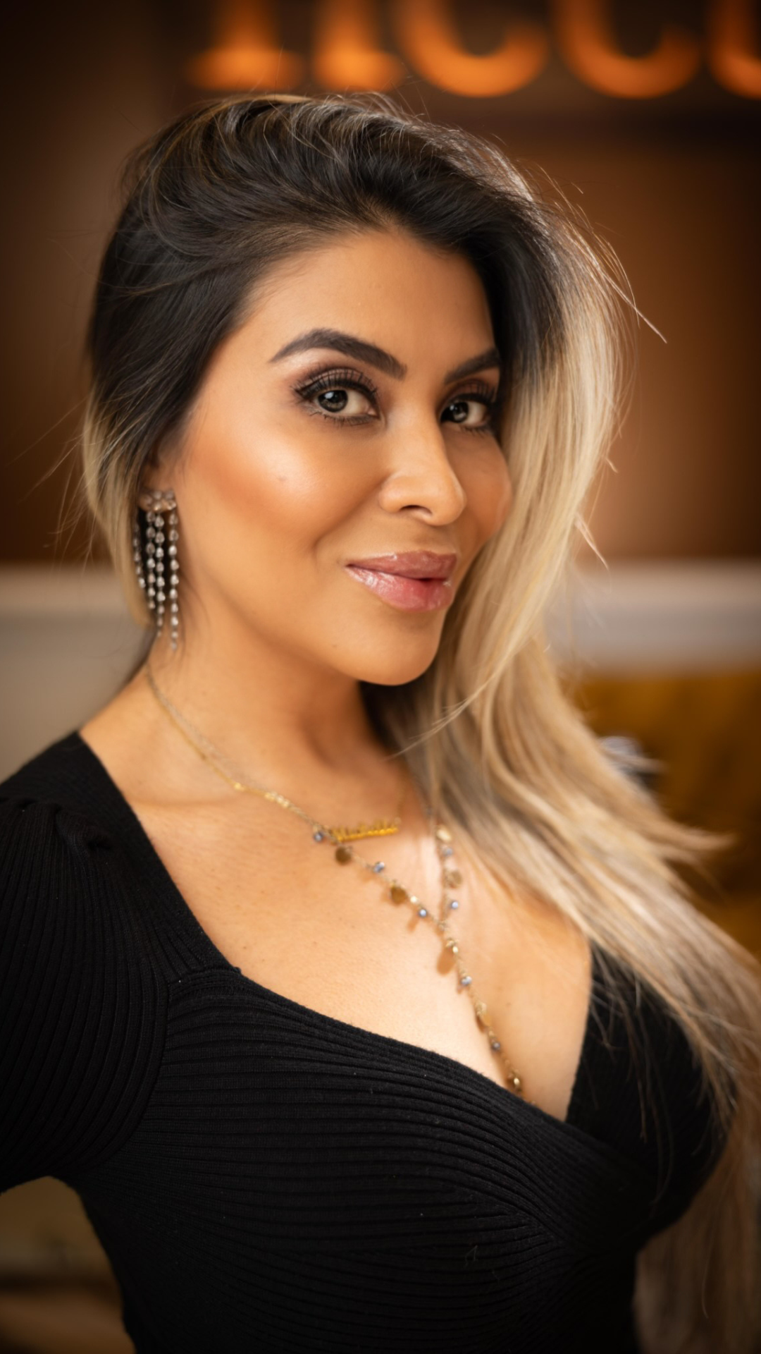 A woman with long blond hair styled over one shoulder, wearing makeup, diamond earrings, a layered necklace with small pendants, and a black top with a deep neckline, smiling softly in an indoor setting.