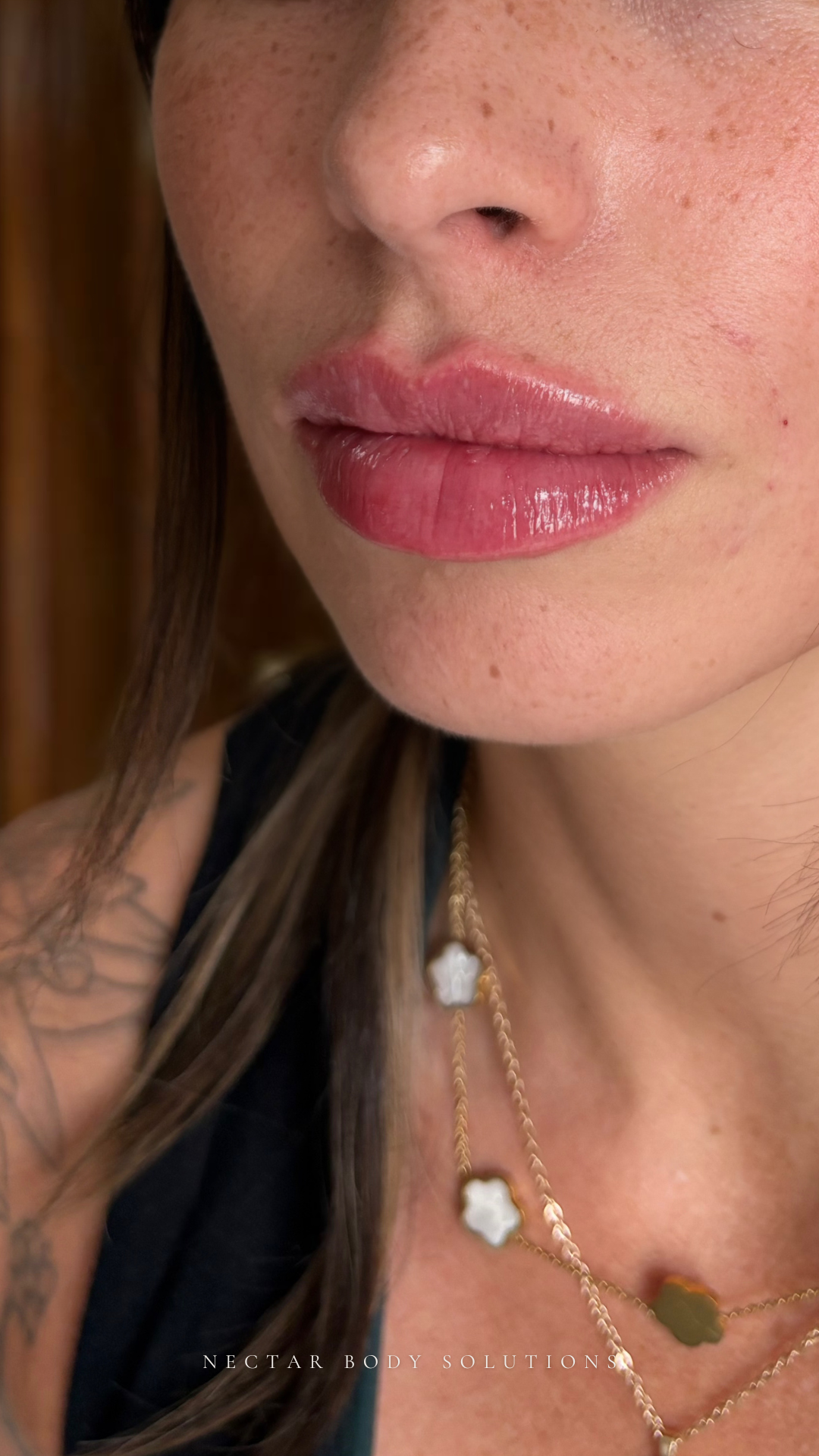 Close-up of a woman's lips with glossy pink lipstick, part of her face, shoulder, and necklaces visible, with tattoo on shoulder.