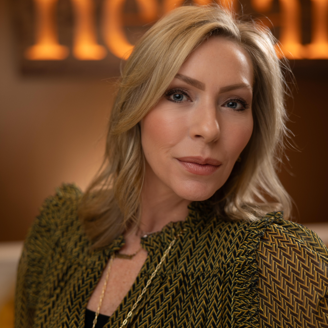 A woman with blonde hair and light skin looking at the camera in a warmly lit indoor setting.