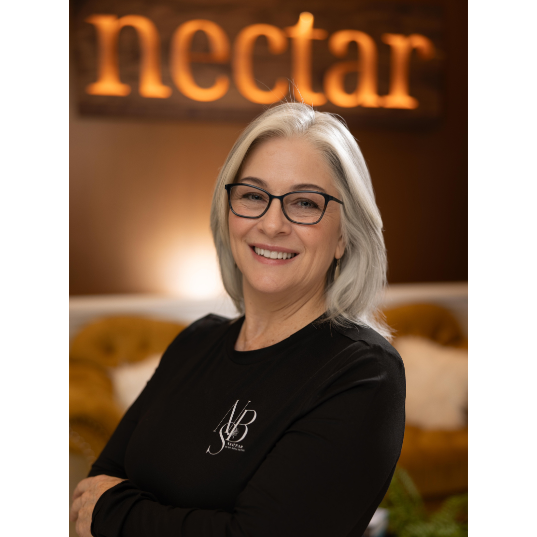 A smiling woman with gray hair and glasses, wearing a black shirt with a logo, standing in front of a blurred background with the word 'nectar' illuminated.