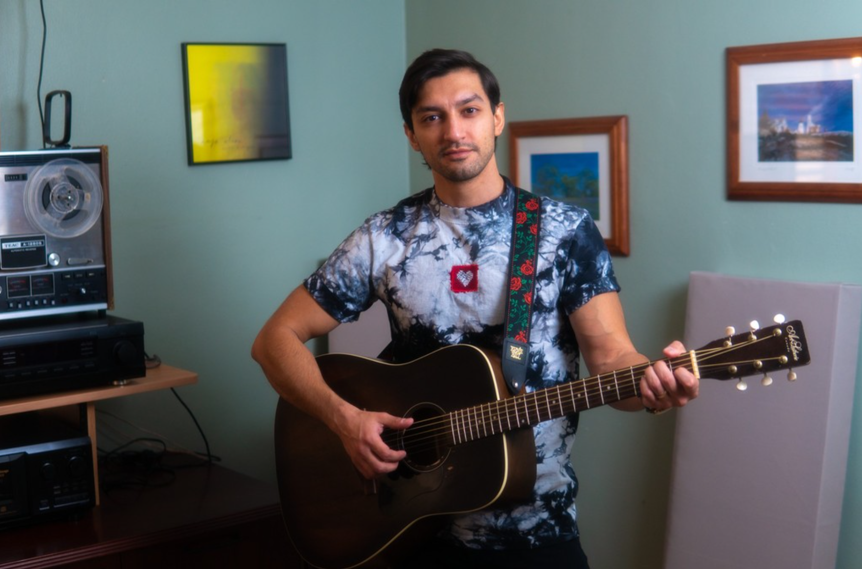 Man playing acoustic guitar indoors with framed pictures on the wall behind him.