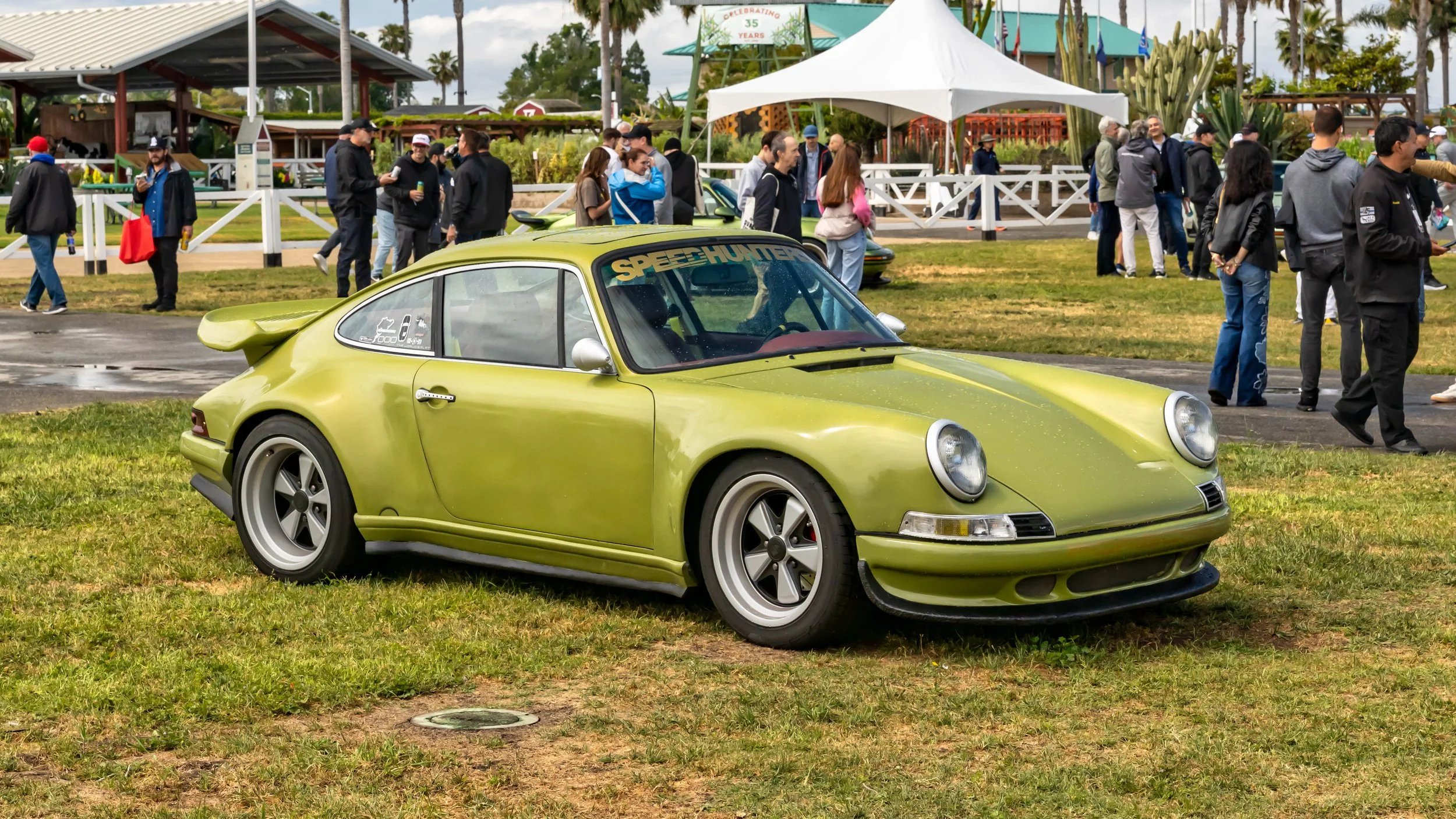 Air | Water 2025 — Orange County Fairgrounds — A heavily modified, lime green classic Porsche 911 is parked on a grassy field at an outdoor car event. The car features a prominent 'ducktail' spoiler, deep-dish Fuchs-style wheels, and a 'Speedhunters'