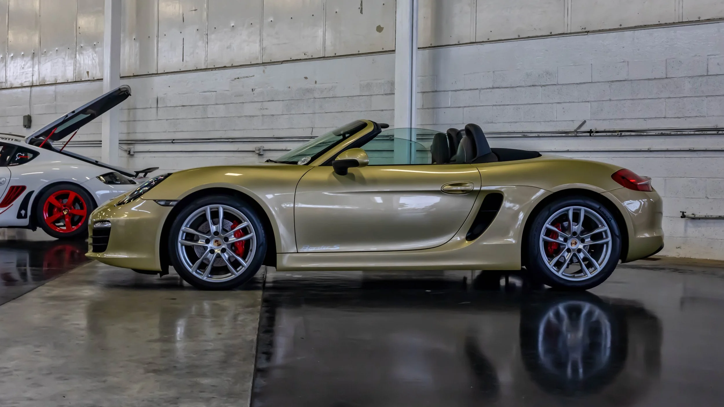Air | Water 2025 — Orange County Fairgrounds — A side profile view of a Lime Gold Metallic Porsche Boxster S (981) with its convertible top down, parked inside a garage or workshop. The car is equipped with 20-inch Carrera S wheels and red brake cali