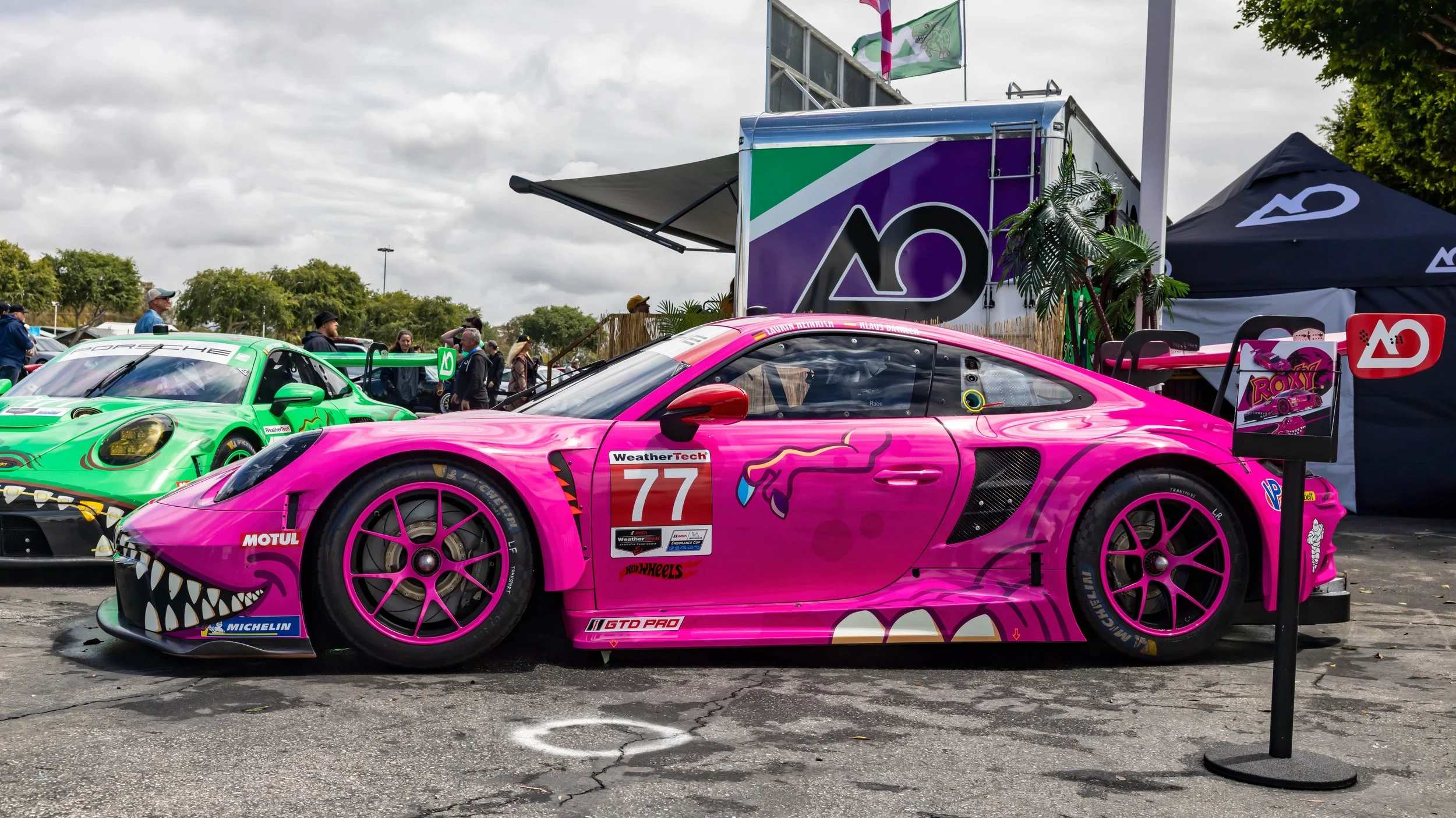 Air | Water 2025 — Orange County Fairgrounds — A side profile view of the AO Racing No. 77 Porsche 911 GT3 R in a vibrant pink 'Roxy' dinosaur livery, parked in a race paddock. The car features matching pink multi-spoke, center-lock wheels, a large s