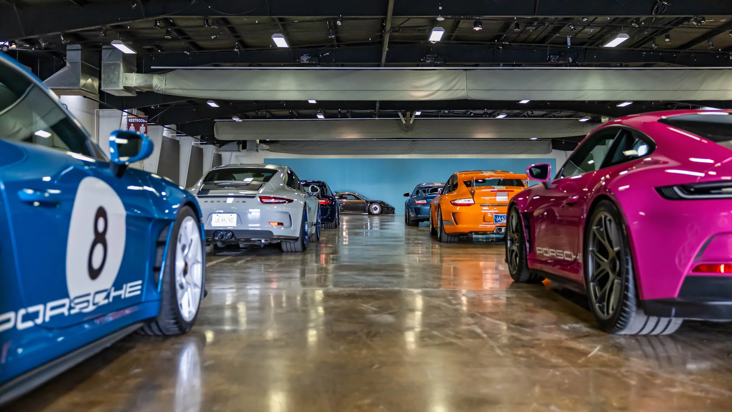 Air | Water 2025 — Orange County Fairgrounds — A low-angle, ground-level shot captures a lineup of colorful Porsche models parked inside a spacious exhibition hall with a polished concrete floor. In the foreground, a blue 991-generation 911 with a wh