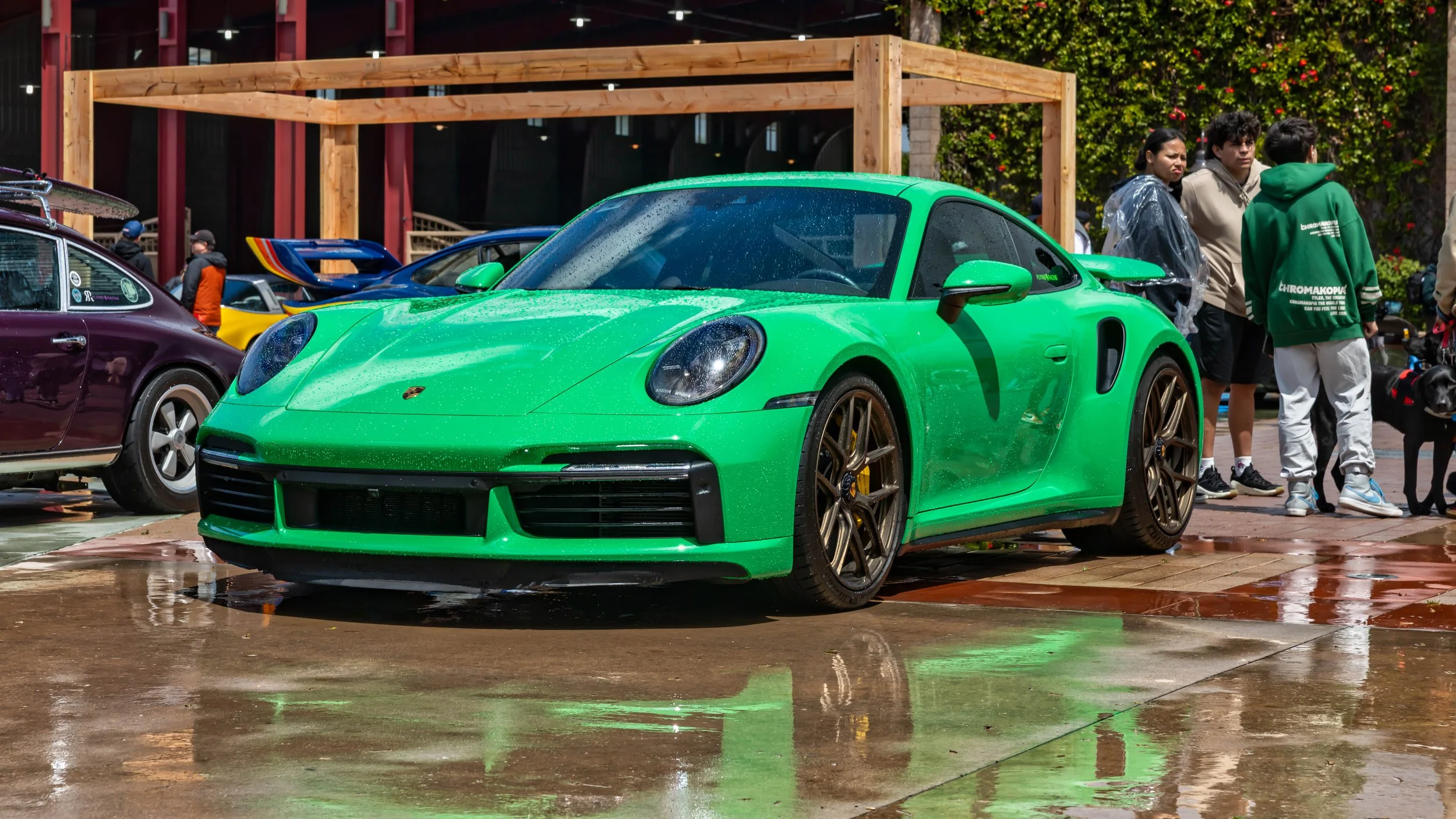 Air | Water 2025 — Orange County Fairgrounds — A vibrant Python Green Porsche 911 Turbo S of the 992 generation is parked at an outdoor car event on wet, reflective pavement. The car is covered in water droplets and features bronze center-lock wheels