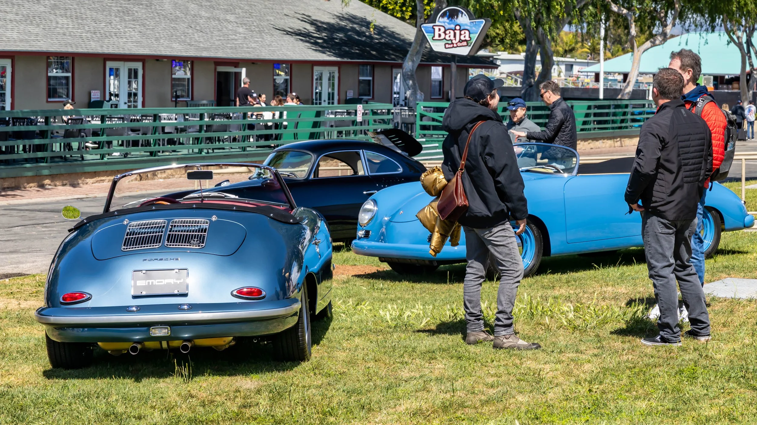 Air | Water 2025 — Emory Motorsports — OC Fairgrounds — Three classic Porsche 356 Emory Outlaws are displayed on a grassy field during a sunny day. In the foreground, a metallic blue 356 Emory convertible is shown from the rear, featuring twin engine