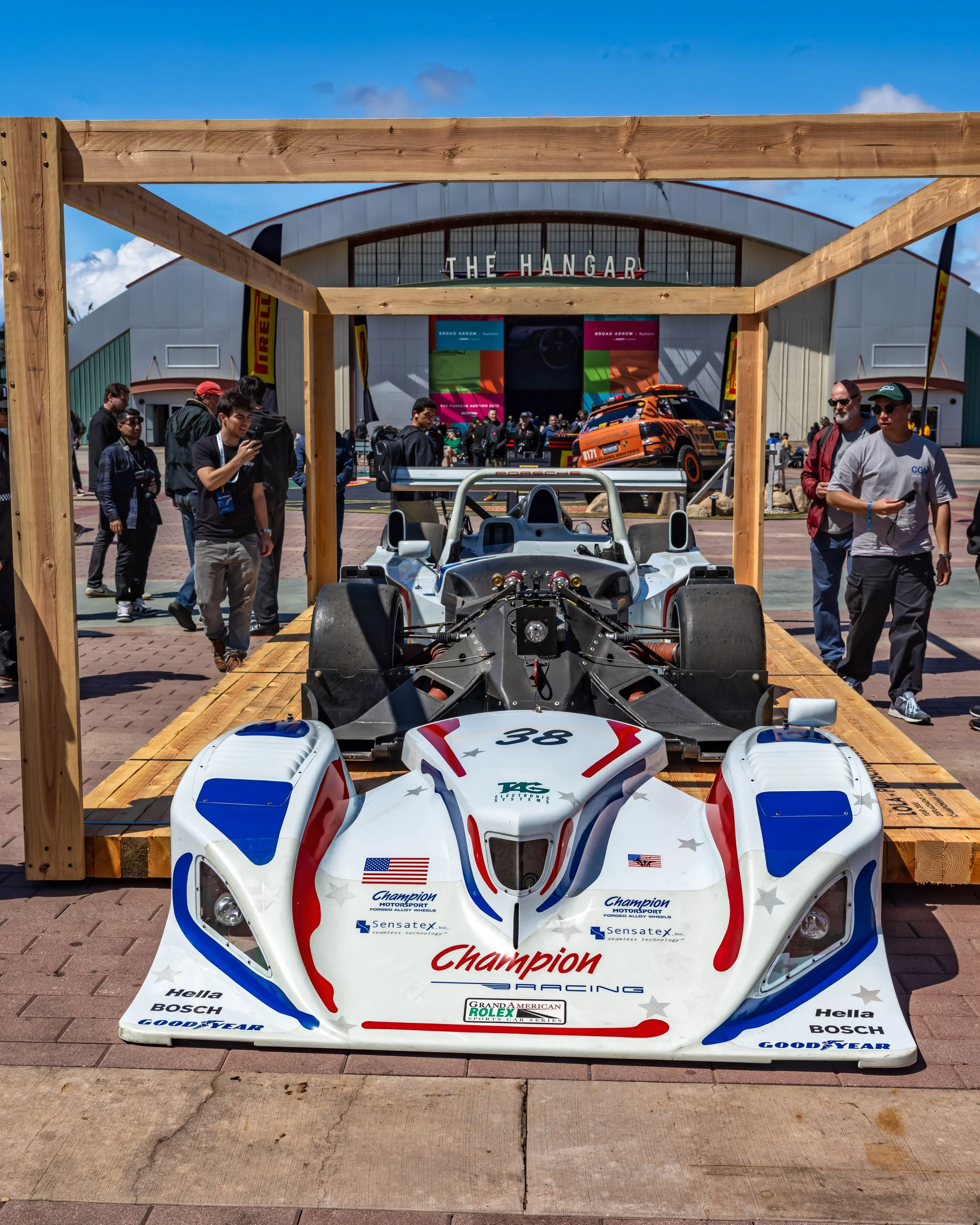 Air | Water 2025 — Orange County Fairgrounds — A front view of a No. 38 Lola B2K/10 race car with a white, red, and blue Champion Racing livery, displayed on a wooden platform. The car is outdoors on a brick patio in front of a large building labeled