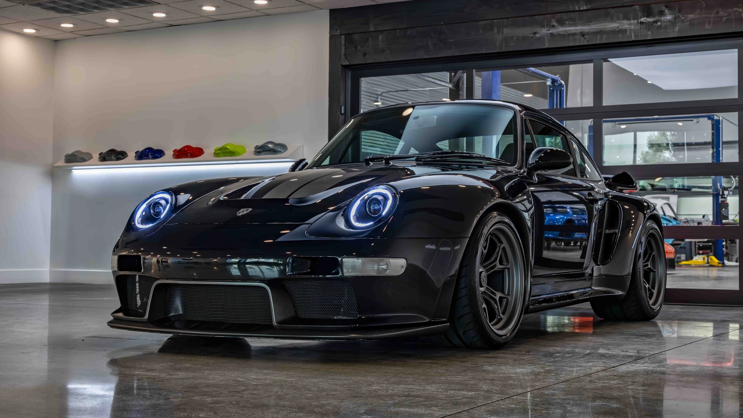Turbo Florio 5 — NVIDIA Black Metallic 1996 Gunther Werks remastered Porsche 911 Turbo (993) parked in the Gunther Werks showroom, left front‑three‑quarter view with exposed carbon-fiber stripe and 18" Magnesium wheels. — #TurboFlorio5
