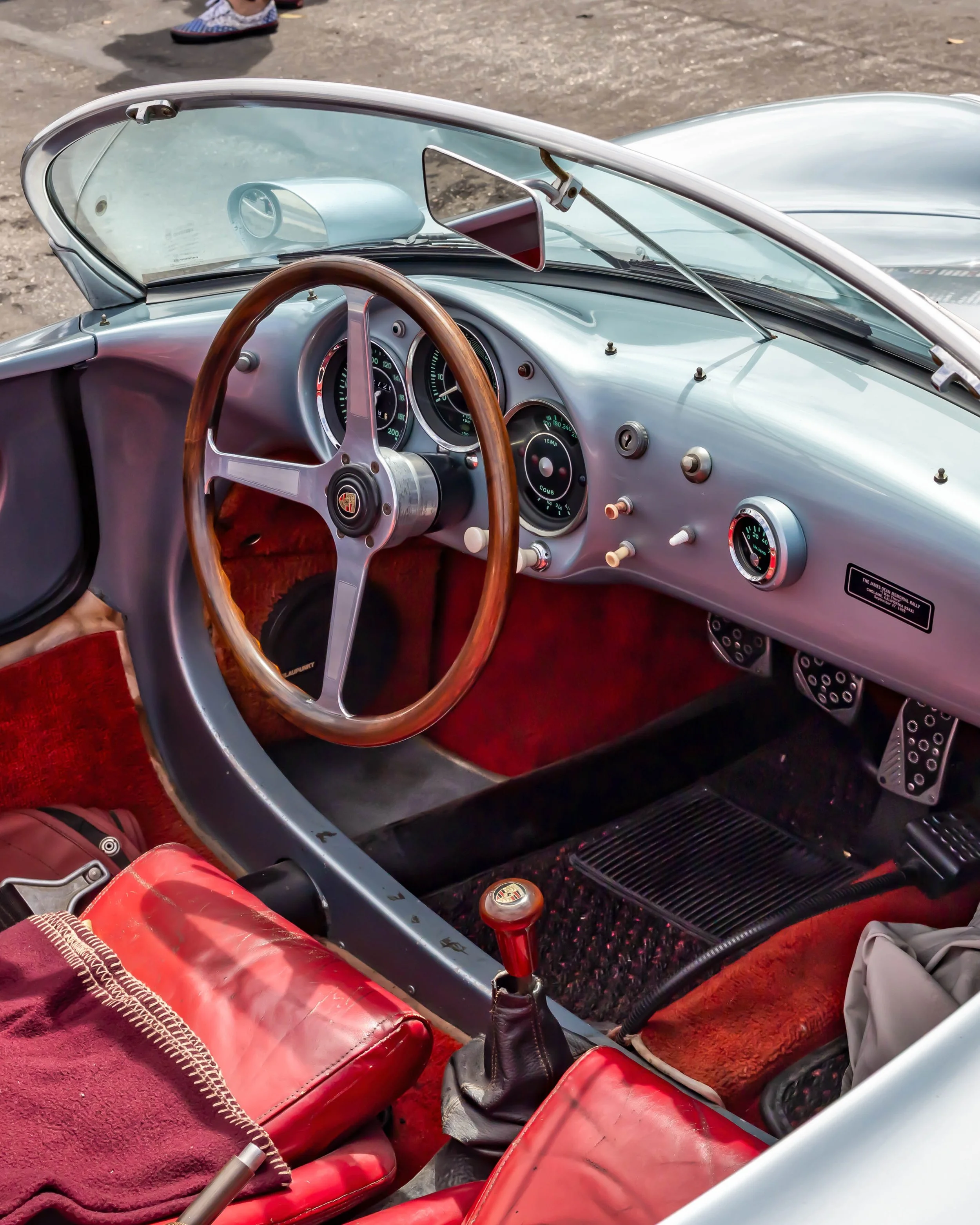 Air | Water 2025 — Orange County Fairgrounds — A high-angle view into the driver's side cockpit of a silver Porsche 550 Spyder replica. The interior features vibrant red leather bucket seats, red carpeting, a wood-rimmed three-spoke steering wheel wi