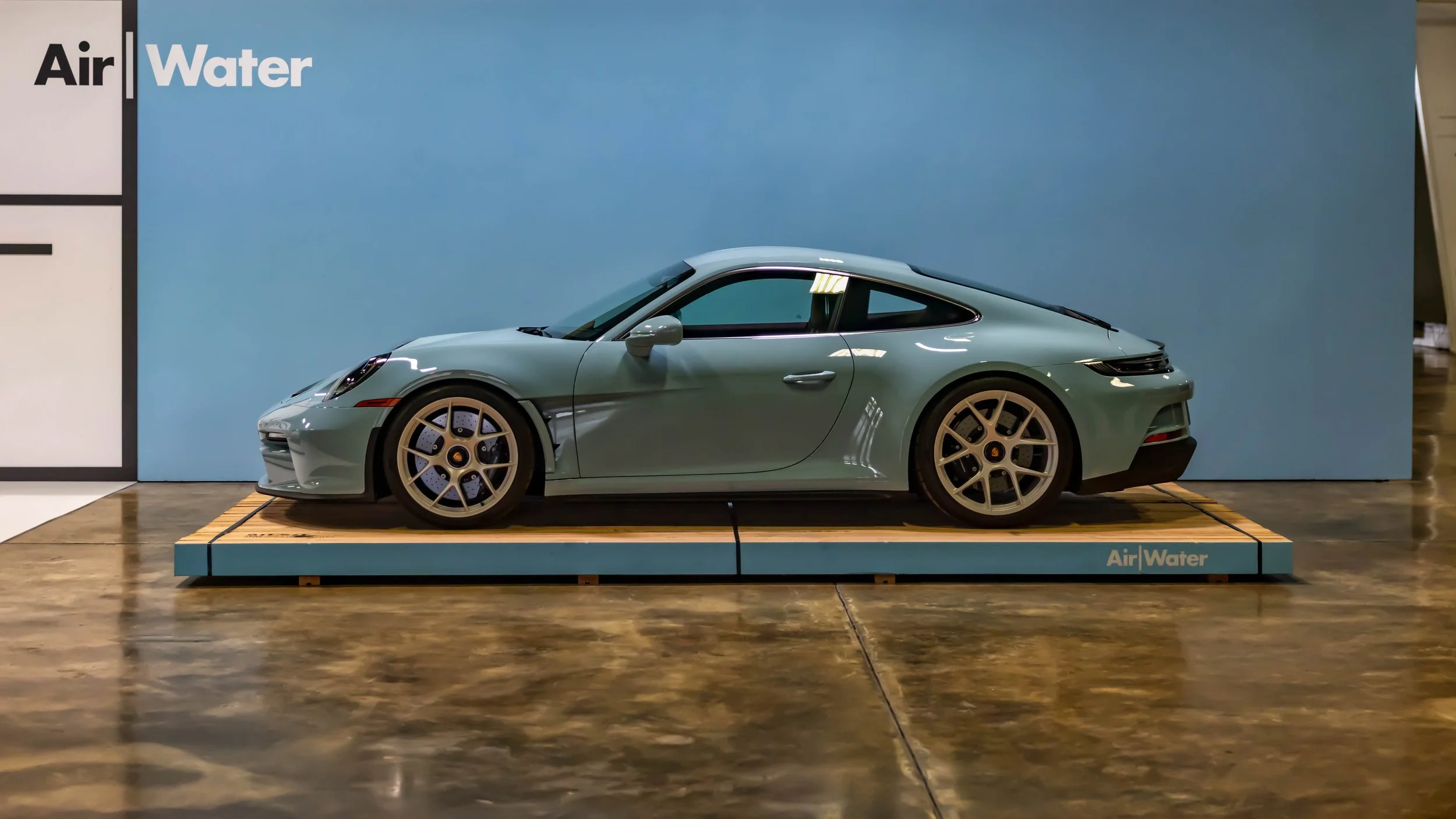 Air | Water 2025 — Orange County Fairgrounds — A light grey-blue 2023 Porsche 911 Sport Classic is displayed in profile on a wooden platform at an indoor event. The car, finished in Sport Grey Metallic, features white Fuchs-style center-lock wheels a