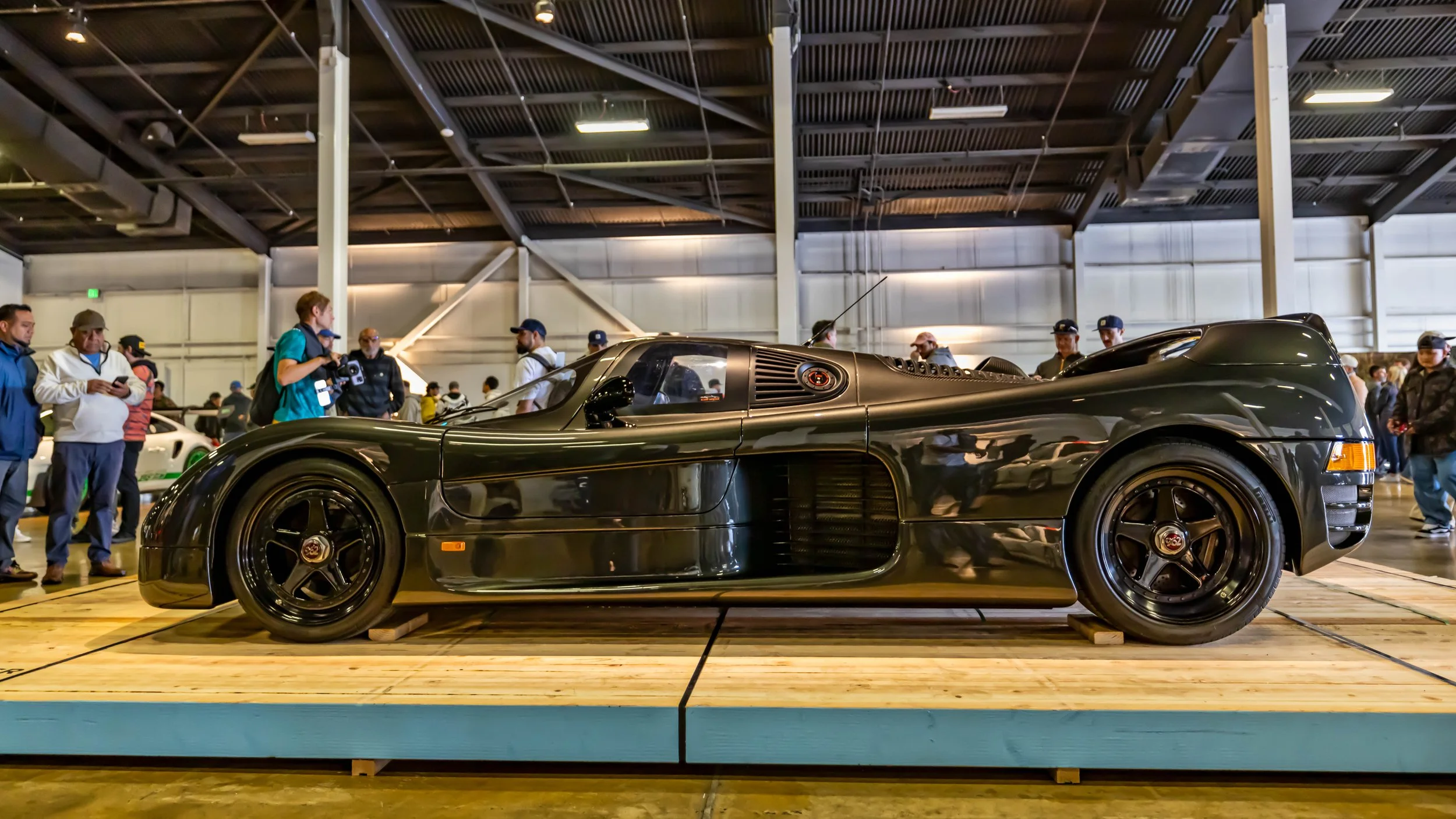 Air | Water 2025 — Orange County Fairgrounds — A dark metallic green Dauer 962 Le Mans, a road-legal version of the Porsche 962 Group C race car, is displayed on a wooden platform at an indoor car show. The car is shown in profile, highlighting its l