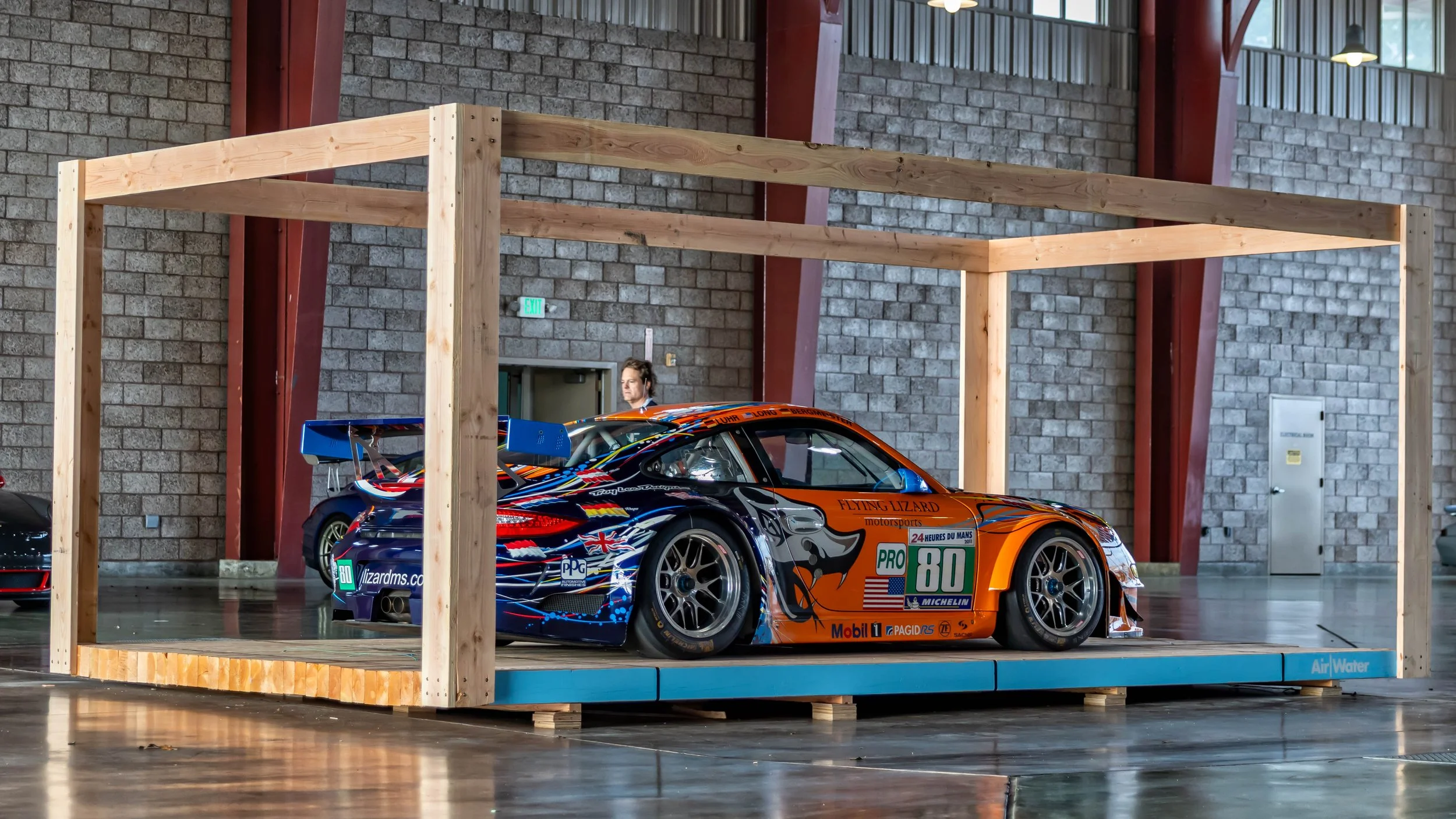 Air | Water 2025 — Orange County Fairgrounds — A 2011 Porsche 911 GT3 RSR from Flying Lizard Motorsports, number 80, is displayed on a raised platform within a large wooden frame. The car features its iconic orange, blue, and black livery from the 20