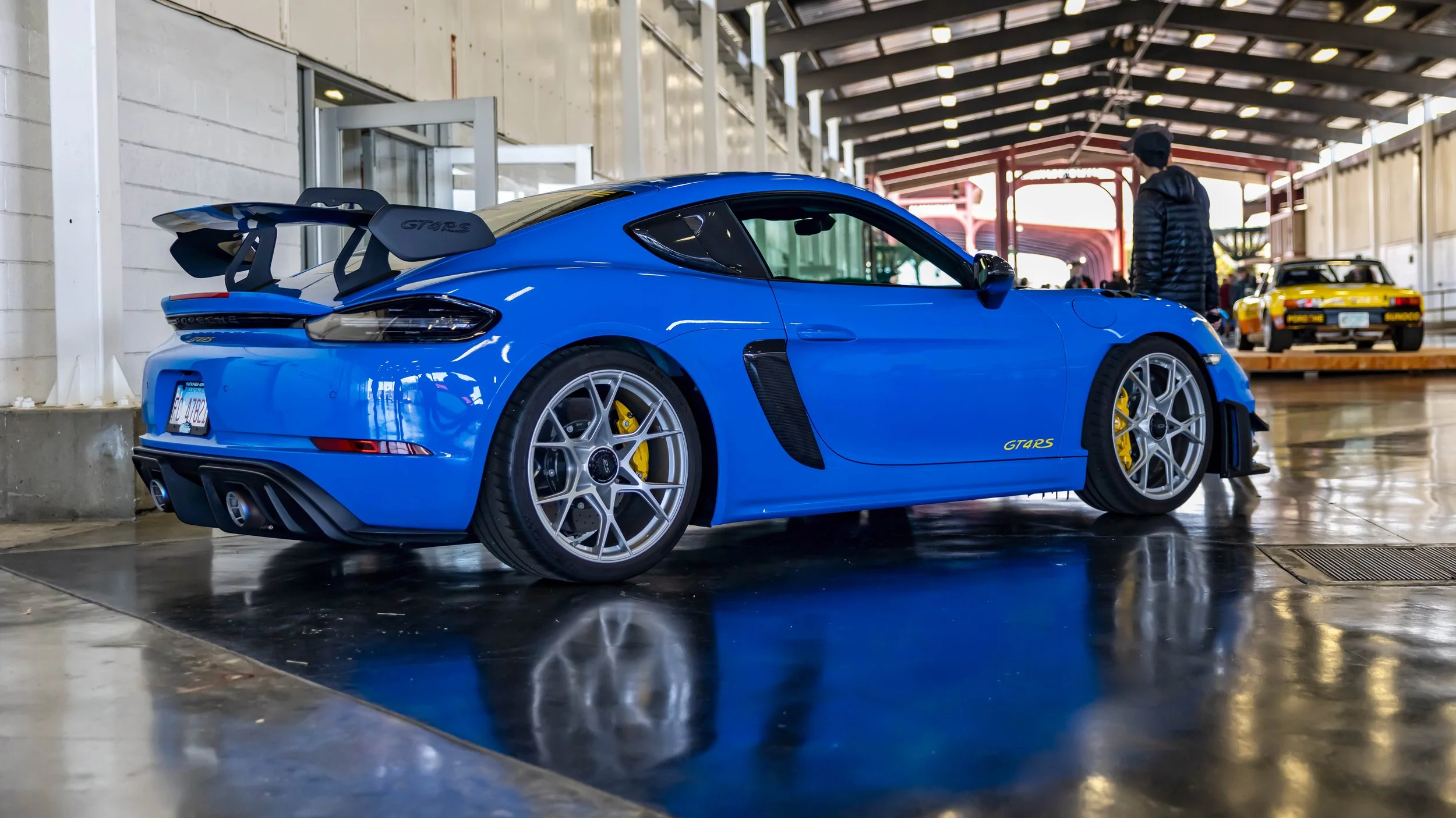 Air | Water 2025 — Orange County Fairgrounds — A low-angle, rear three-quarter view of a Shark Blue Porsche 718 Cayman GT4 RS parked inside a well-lit industrial garage or event space. The car's reflection is clearly visible on the dark, glossy floor