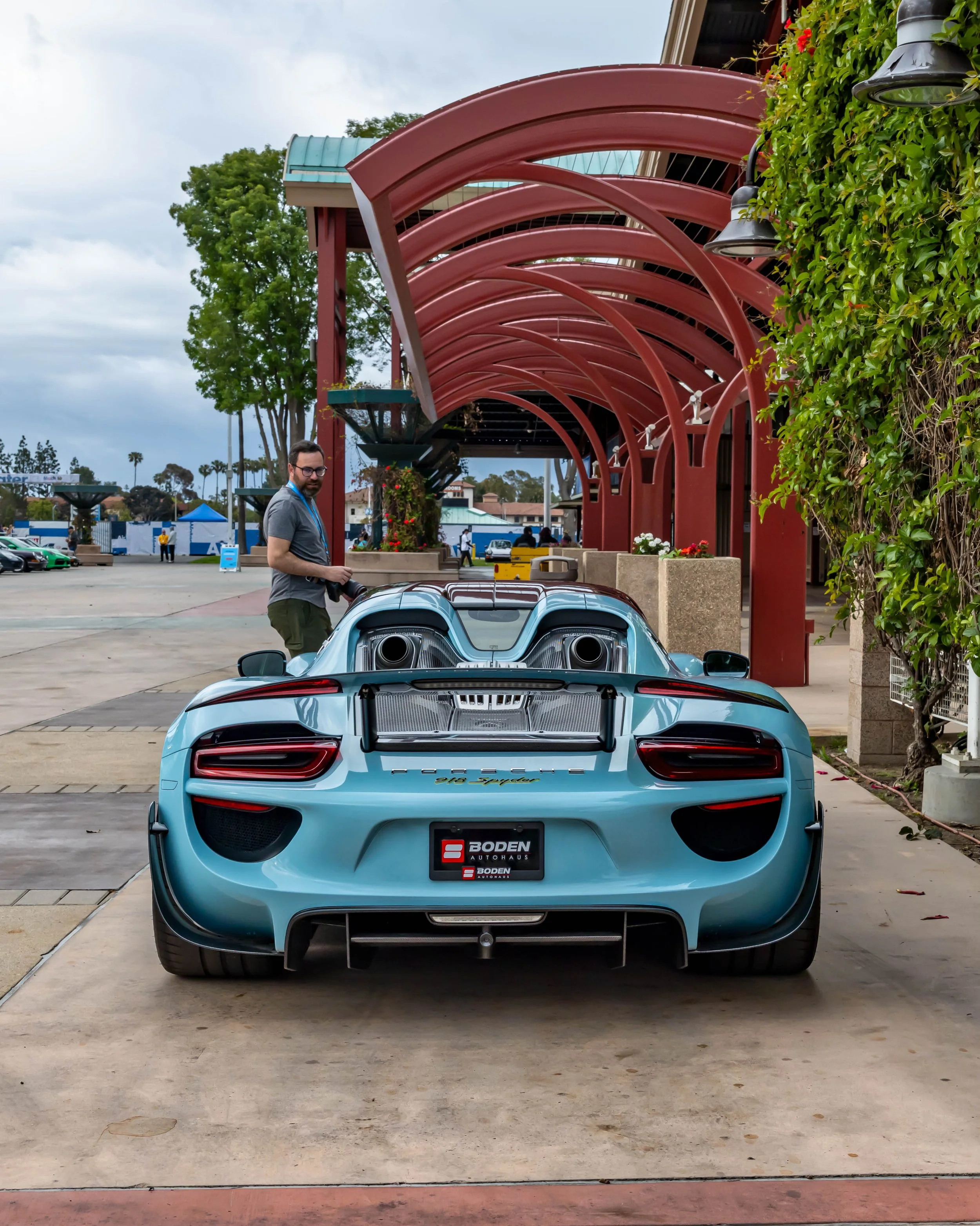 Air | Water 2025 — Orange County Fairgrounds — A light blue Porsche 918 Spyder is parked on a concrete walkway under a distinctive red arched awning. The car is viewed from the rear, highlighting its top-mounted exhaust pipes, active rear spoiler, an