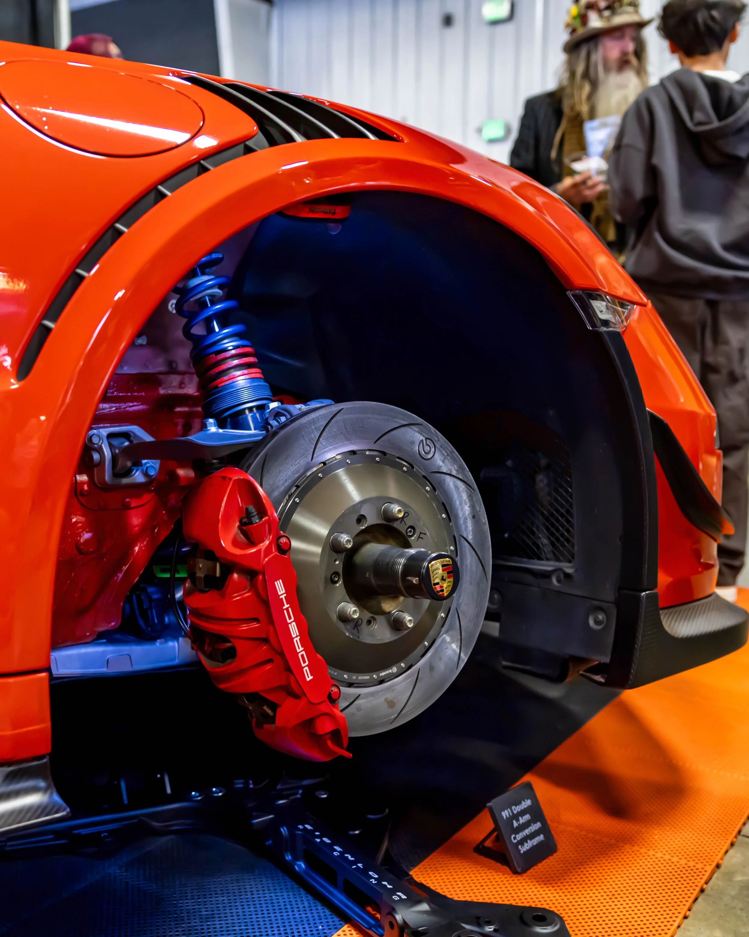 Air | Water 2025 — Orange County Fairgrounds — A detailed close-up of the front driver's side of an orange Porsche 991, displayed with the wheel removed to showcase its Eisenlohr Racing suspension and braking system. The view highlights a large red P