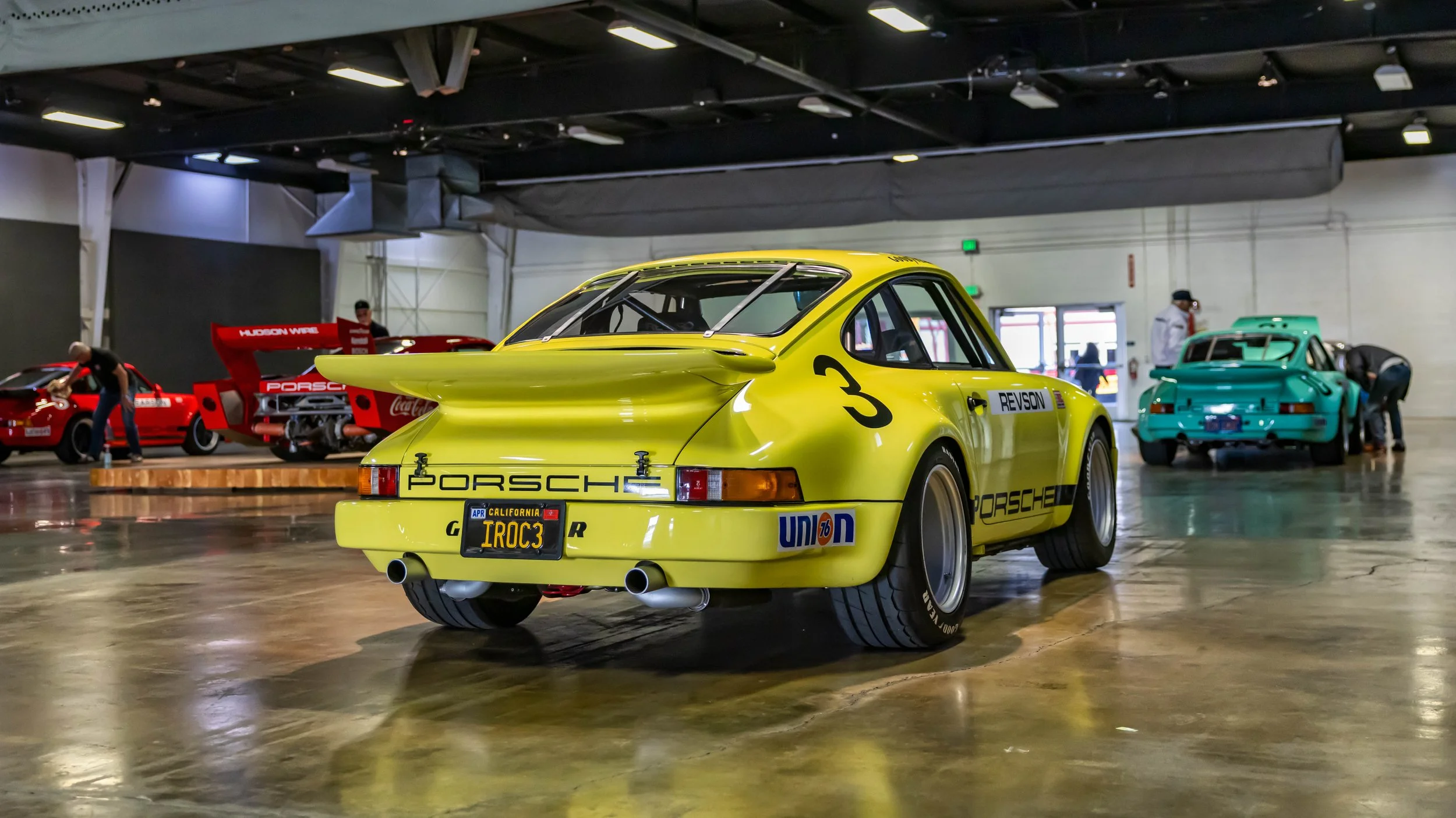 Air | Water 2025 — Orange County Fairgrounds — A low-angle, rear three-quarter view of a yellow 1974 Porsche 911 Carrera RSR IROC race car parked inside a large garage or showroom. The car, featuring the number '3' and 'REVSON' livery, is displayed o