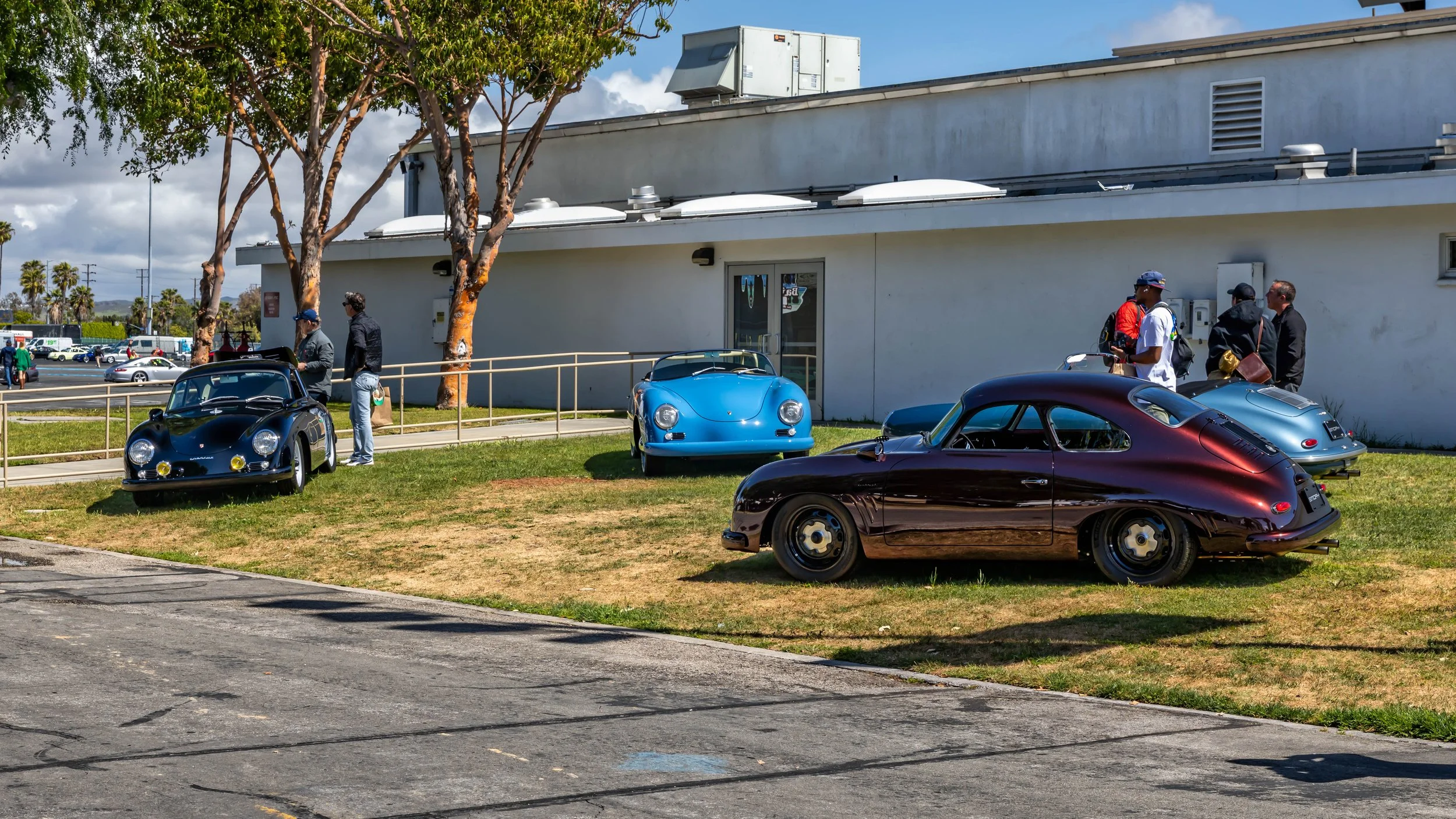 Air | Water 2025 — Emory Motorsports — OC Fairgrounds — A custom Superior Red Metallic Porsche 356 Emory Outlaw Special coupe with black steel wheels is parked on a grassy lawn, positioned as the primary subject. In the background, other classic Pors