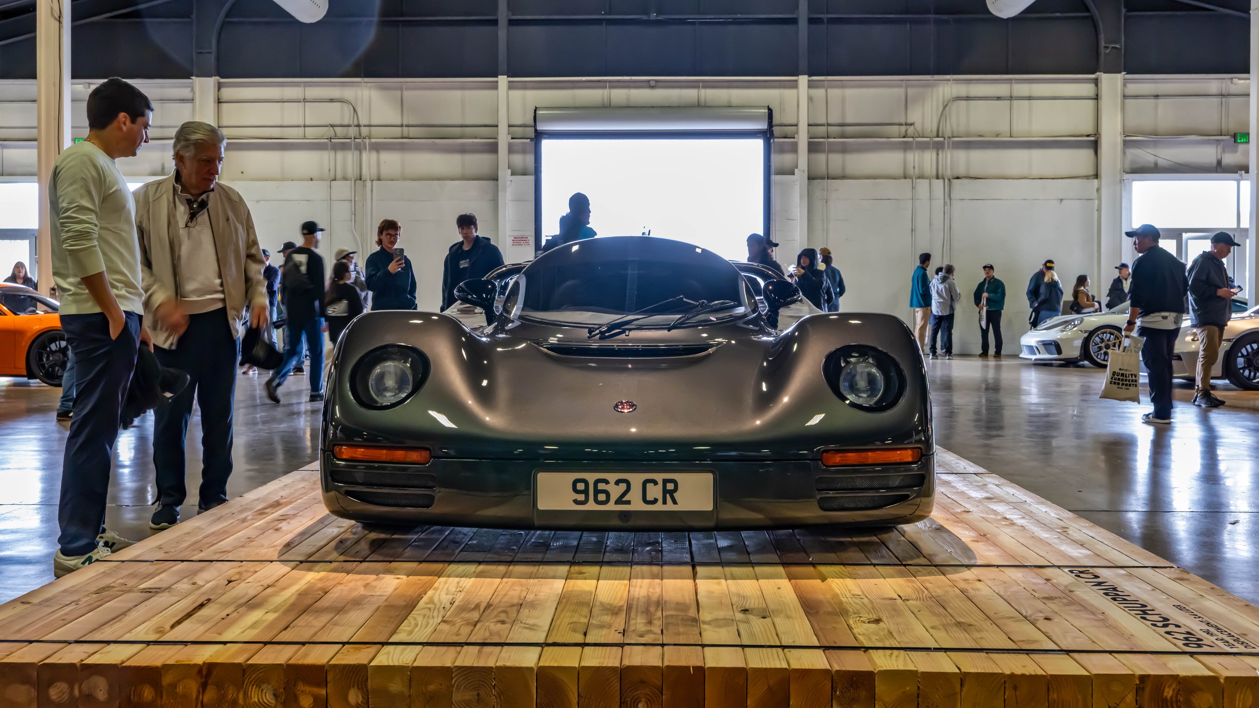 Air | Water 2025 — Orange County Fairgrounds — A low-angle, head-on shot of a rare, gunmetal grey Schuppan 962CR supercar, centrally positioned on a large wooden platform display. The car is the main attraction at an indoor car show held in a exhibit