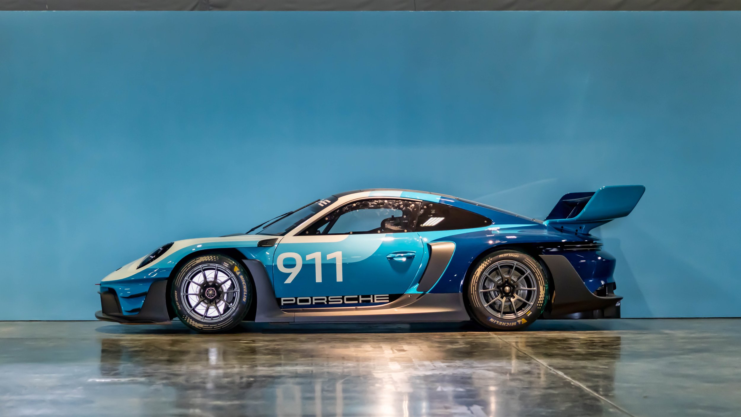 Air | Water 2025 — Orange County Fairgrounds — A side profile view of a Porsche 911 GT2 RS Clubsport 25 track car parked indoors on a reflective, polished concrete floor against a solid light blue wall. The car features a multi-tone blue and white ra