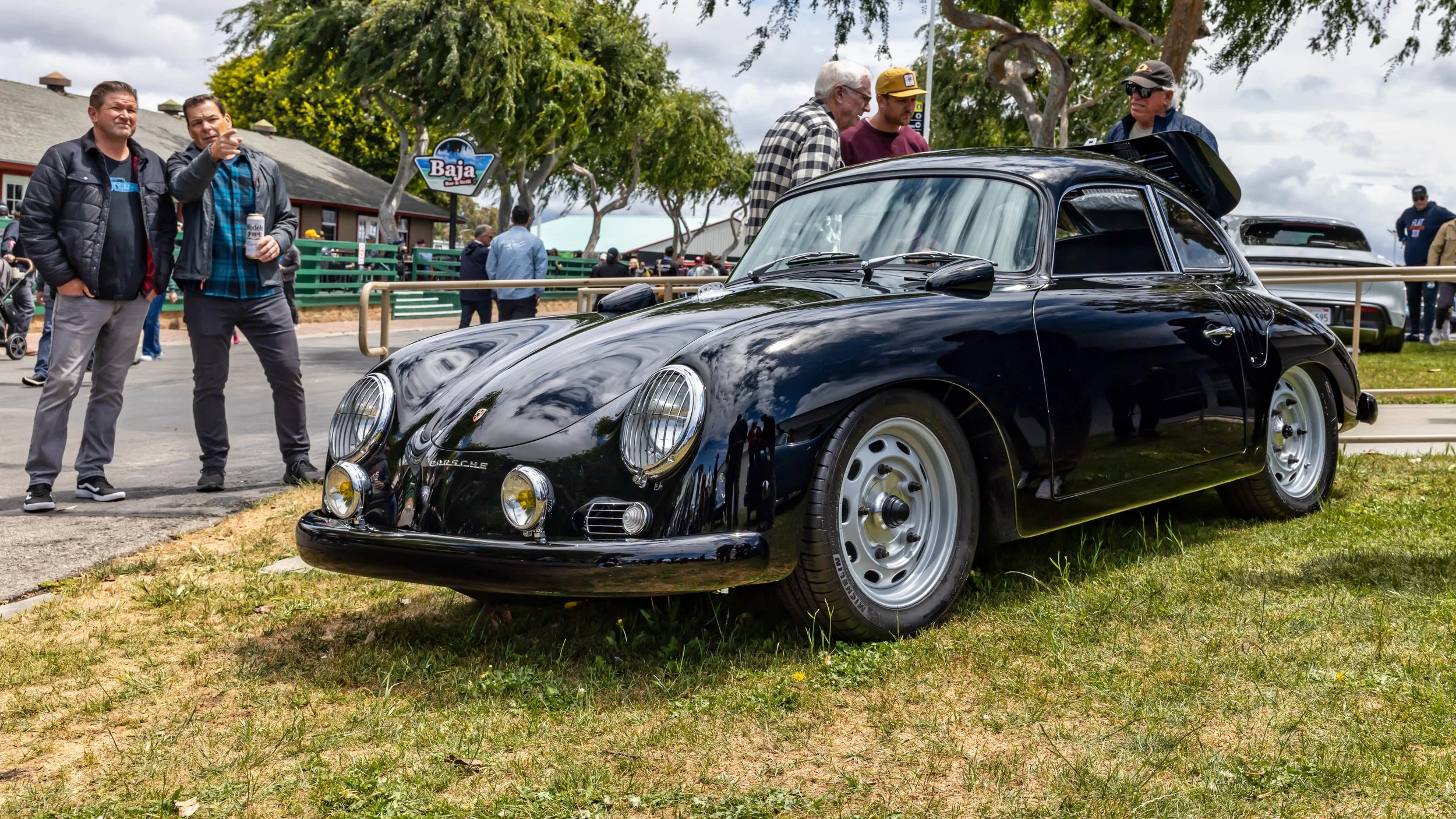 Air | Water 2025 — Emory Motorsports — OC Fairgrounds — A classic, glossy black Porsche 356 Emory Outlaw coupe is parked on a grassy area at an outdoor car show. The car features a lowered stance, silver steel-style wheels, and two auxiliary driving 