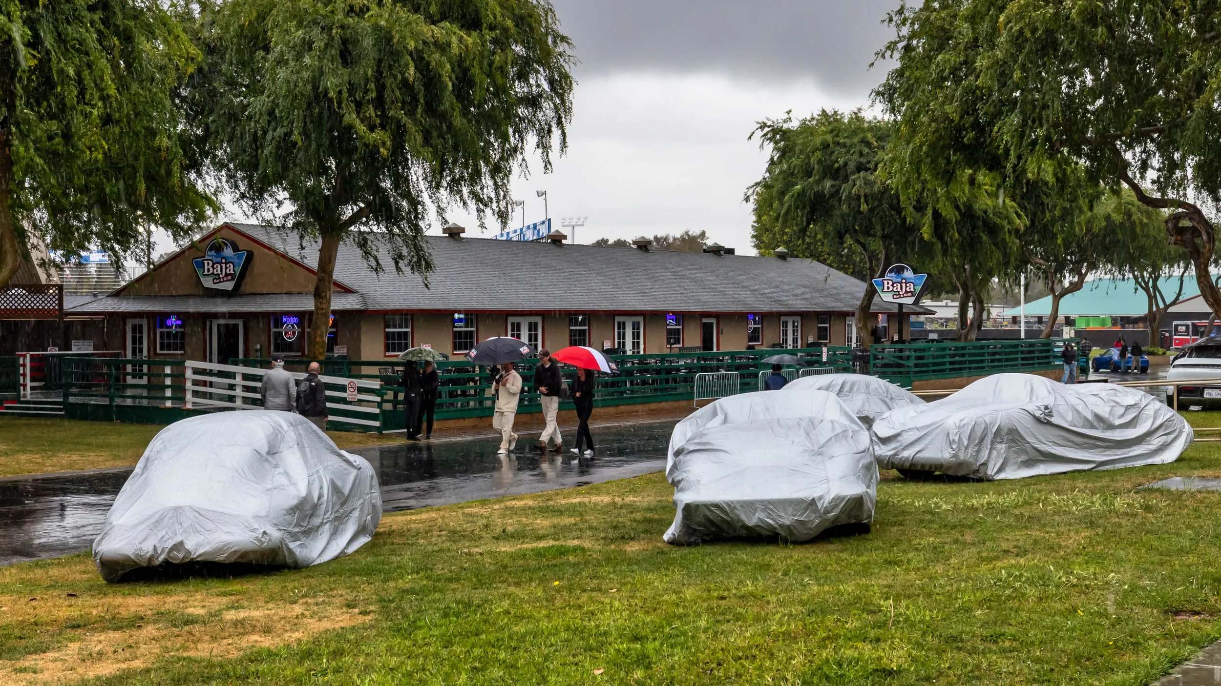 Air | Water 2025 — Emory Motorsports — OC Fairgrounds — Several Emory 356s concealed under silver car covers, are parked on a wet grassy field during a rainy day. In the background, people with umbrellas walk past the "Baja Bar & Grill" building. The