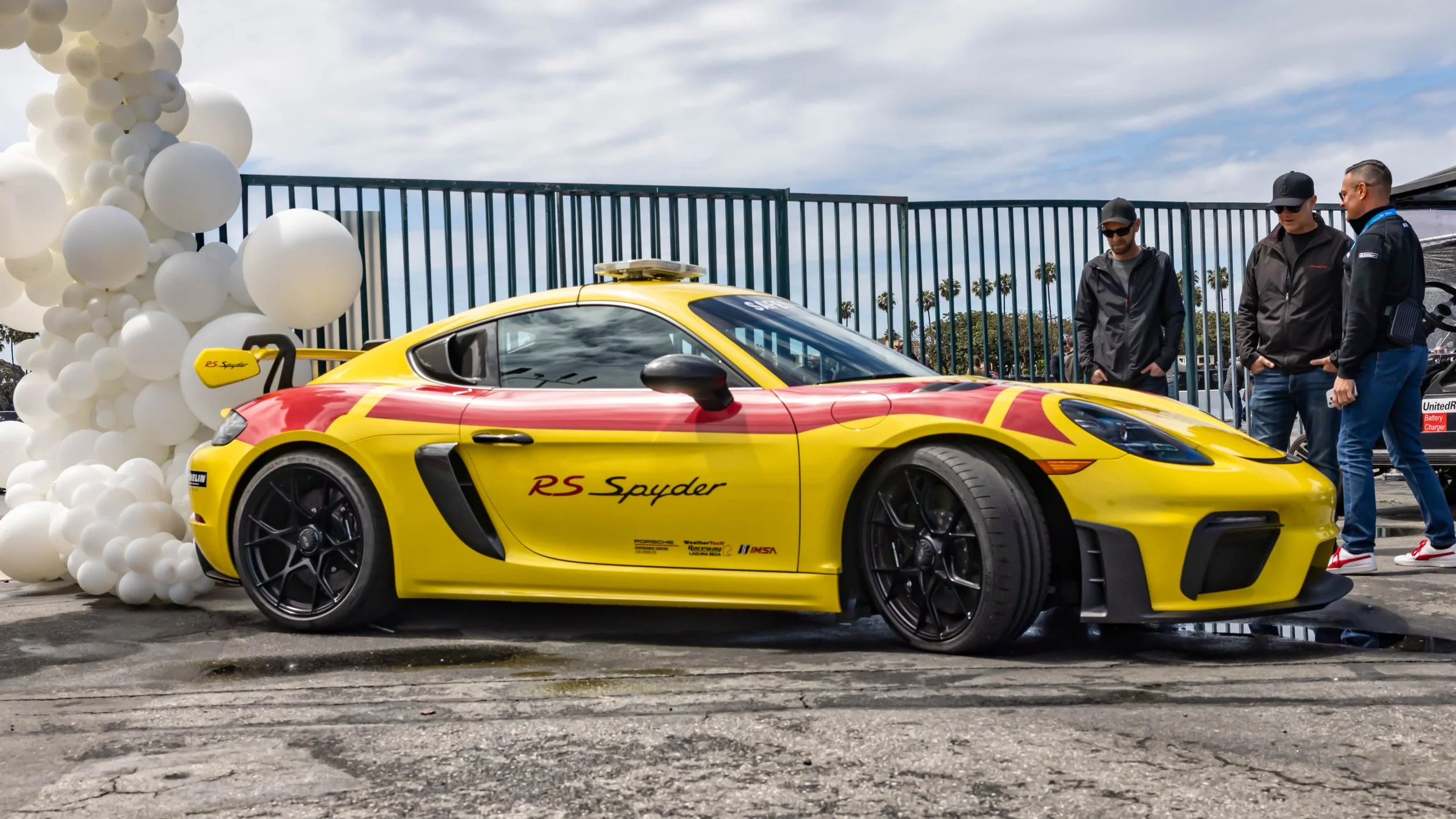 Air | Water 2025 — Orange County Fairgrounds — A low-angle profile view of a yellow Porsche 718 Cayman GT4 RS with a red livery, a tribute to the historic RS Spyder race car. The car is equipped with a roof-mounted light bar, black multi-spoke wheels