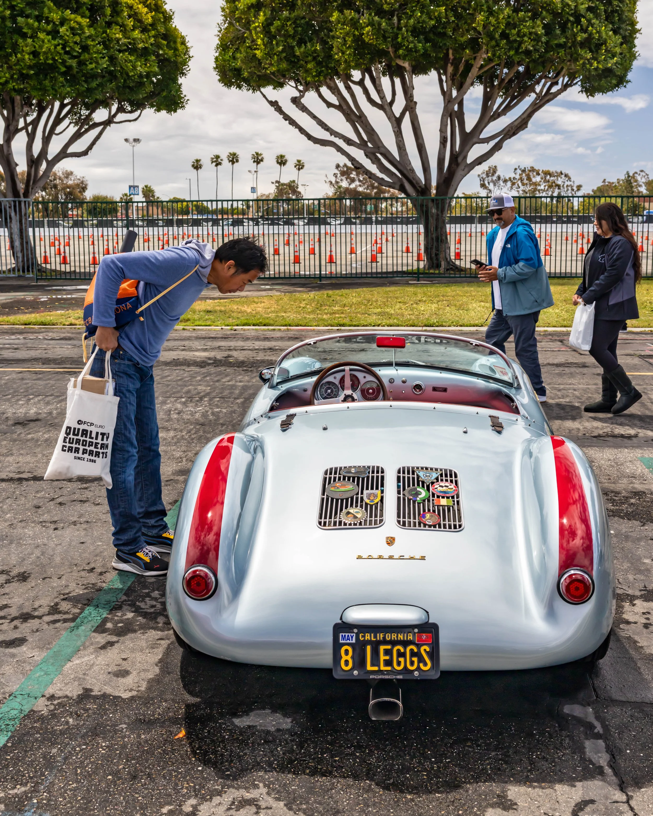 Air | Water 2025 — Orange County Fairgrounds — A rear three-quarter view of a silver Porsche 550 Spyder replica at an outdoor car gathering. A man leans over to inspect the red interior, while other attendees walk by. The car features red stripes ove