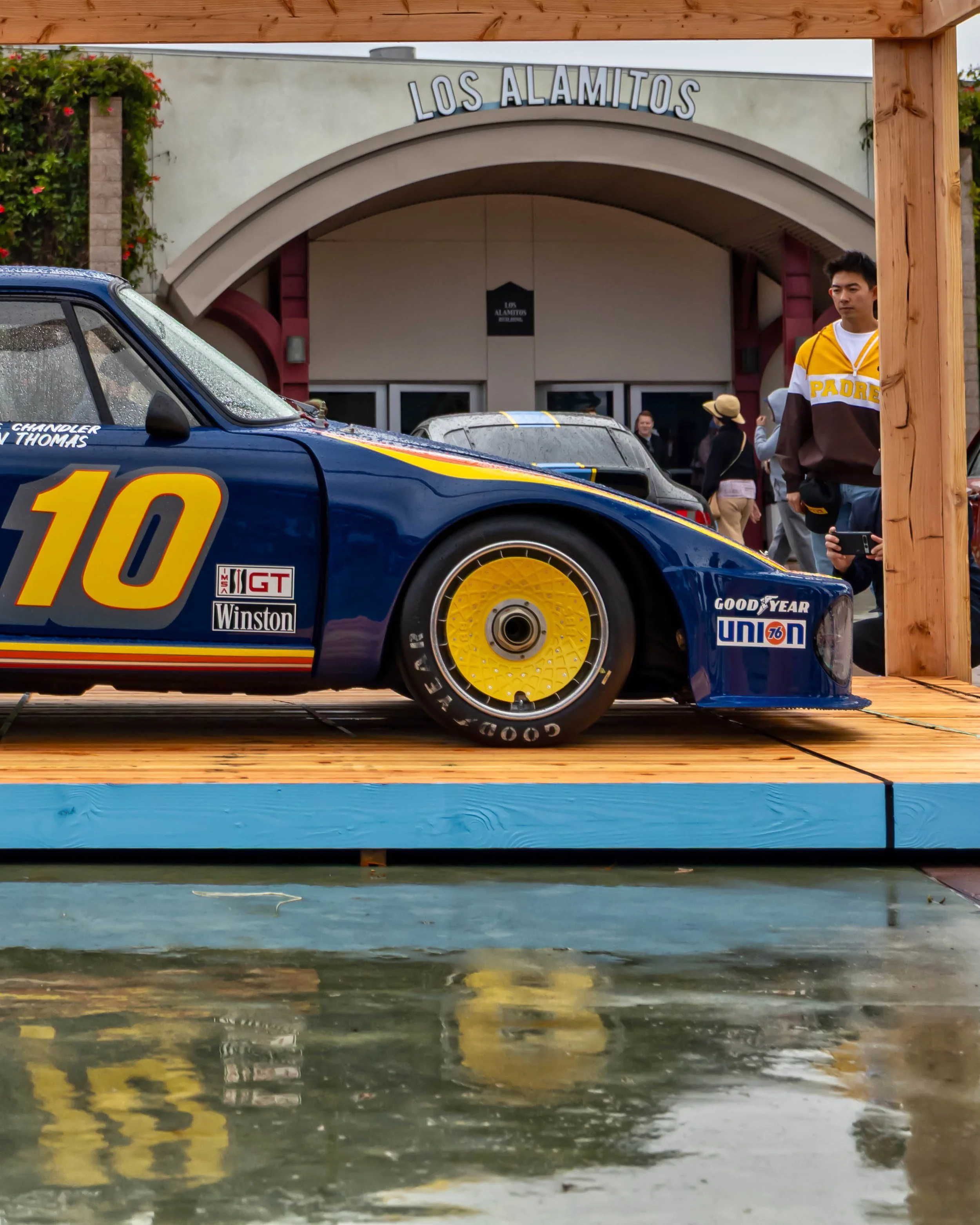 Air | Water 2025 — Orange County Fairgrounds — A low-angle side profile of a vintage blue #10 Interscope Racing Porsche 935 on display at an outdoor car event . The race car, featuring a widebody kit and yellow, orange, and red stripes, is parked on 