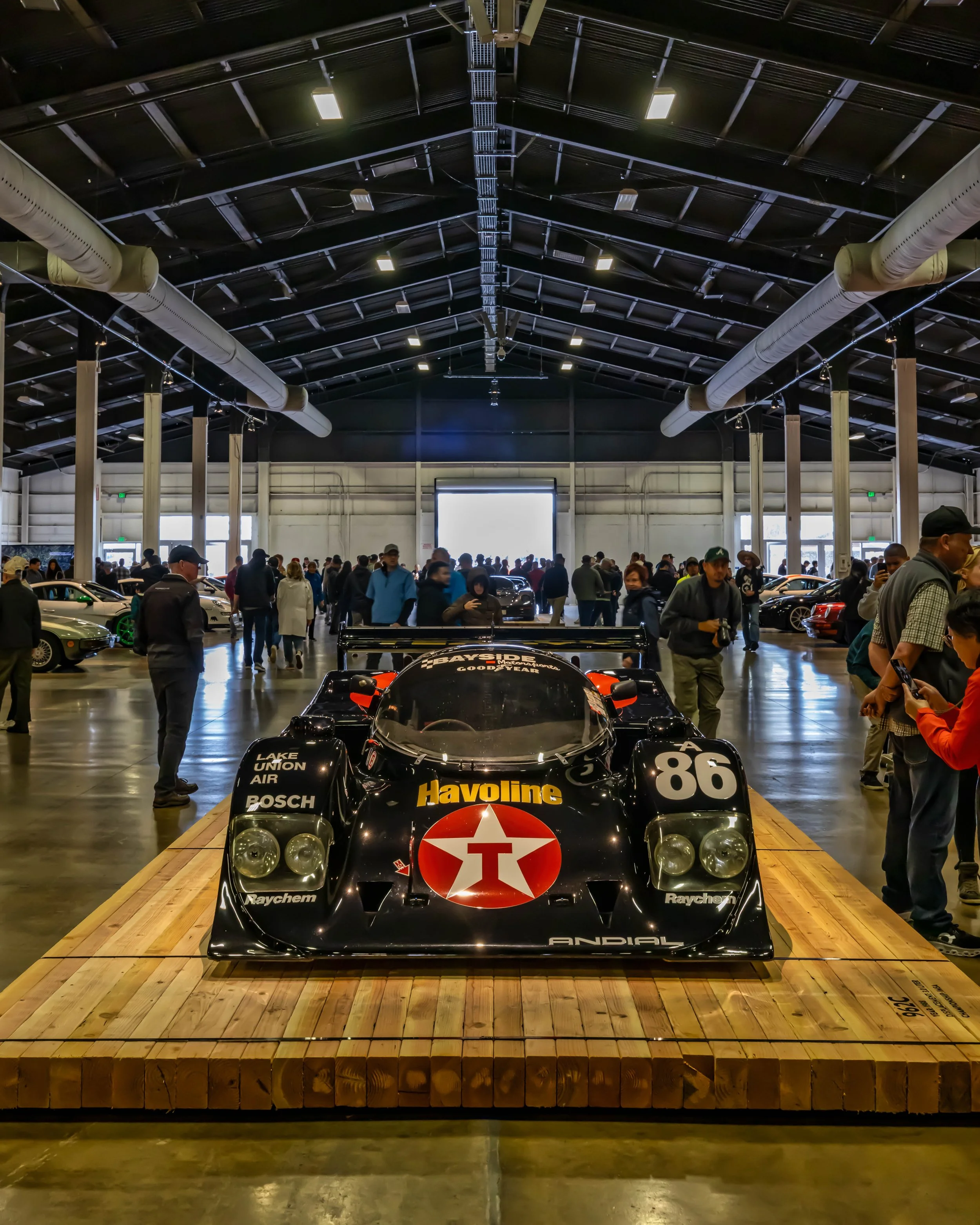 Air | Water 2025 — Orange County Fairgrounds — A head-on view of a black Porsche 962 race car, number 86, with a prominent Havoline/Texaco livery. The car is a central exhibit, elevated on a heavy wooden platform inside a large, crowded exhibition ha
