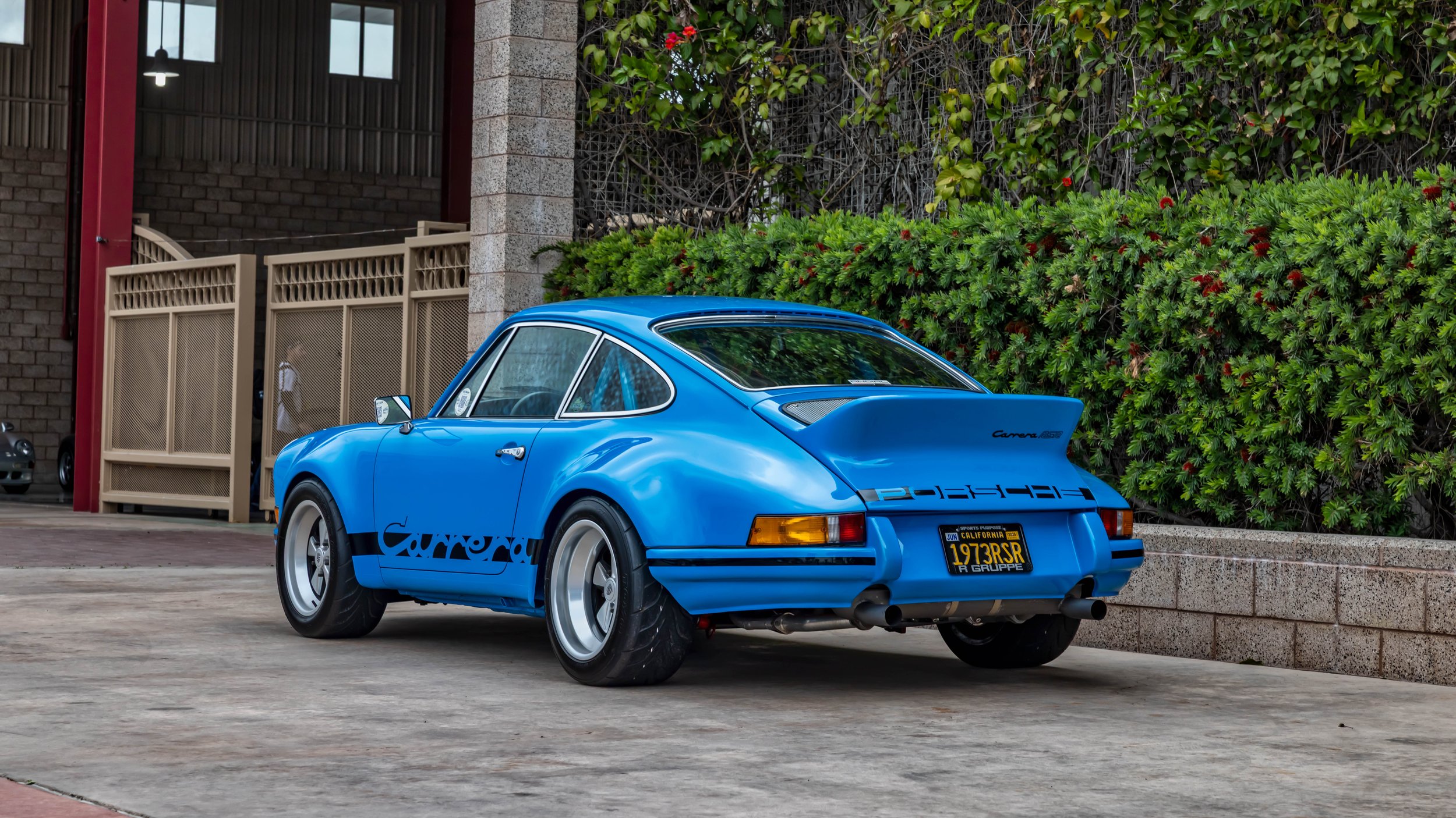 Air | Water 2025 — Orange County Fairgrounds — A light blue 1973 Porsche 911 Carrera RSR is parked on a concrete pavilion, viewed from the rear three-quarter angle. The car features wide fender flares, a prominent ducktail spoiler with 'Carrera RSR' 