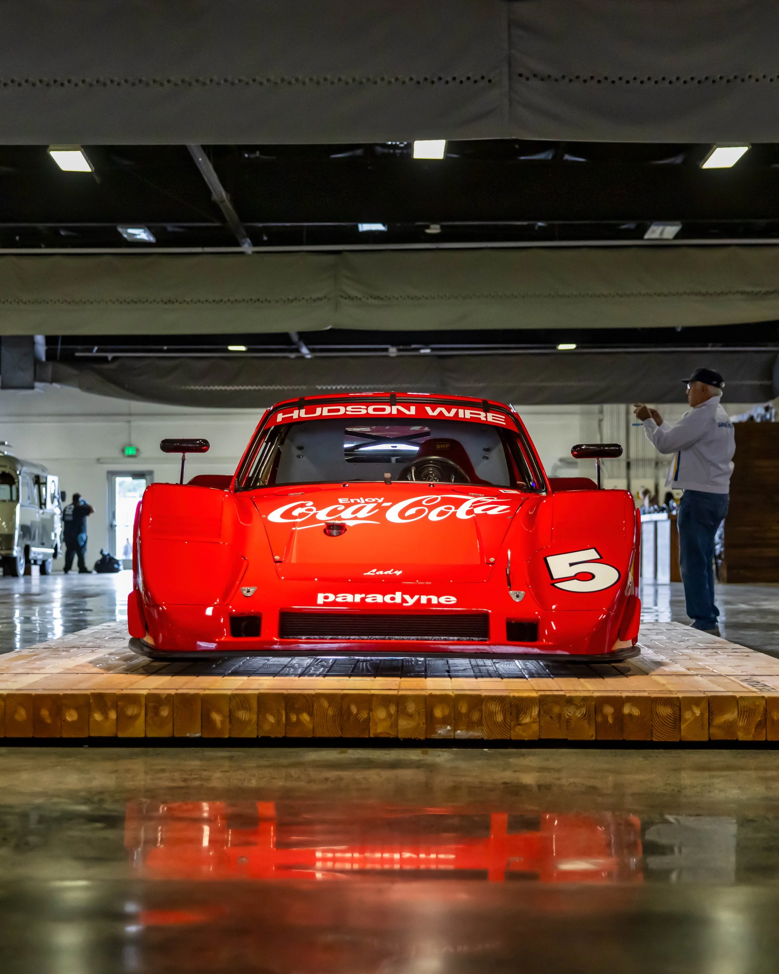 Air | Water 2025 — Orange County Fairgrounds — A low, head-on photograph of the iconic red #5 Coca-Cola Porsche 962 race car, presented on a heavy wooden block platform inside a large exhibition hall. The car's vibrant livery and sponsors, including 