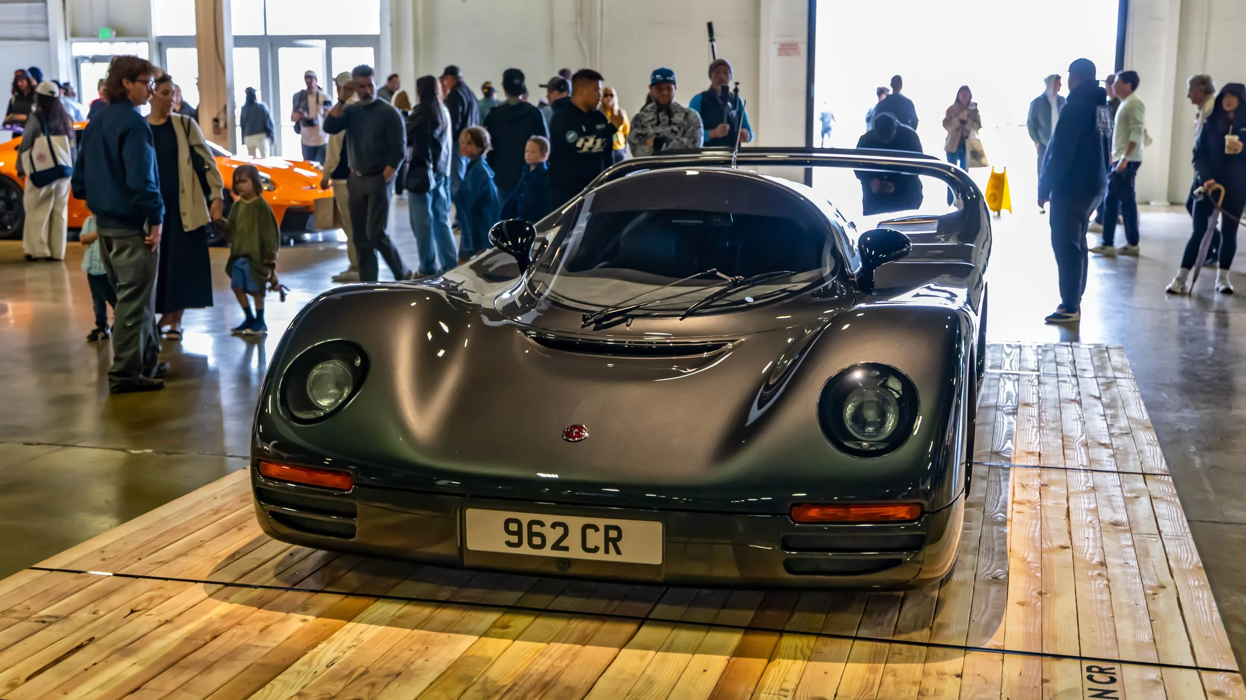 Air | Water 2025 — Orange County Fairgrounds — A low-angle, front three-quarters view of a rare, gunmetal grey Schuppan 962CR supercar. The vehicle, inspired by Le Mans race cars, is showcased on a wooden pallet-like platform inside a brightly lit ex