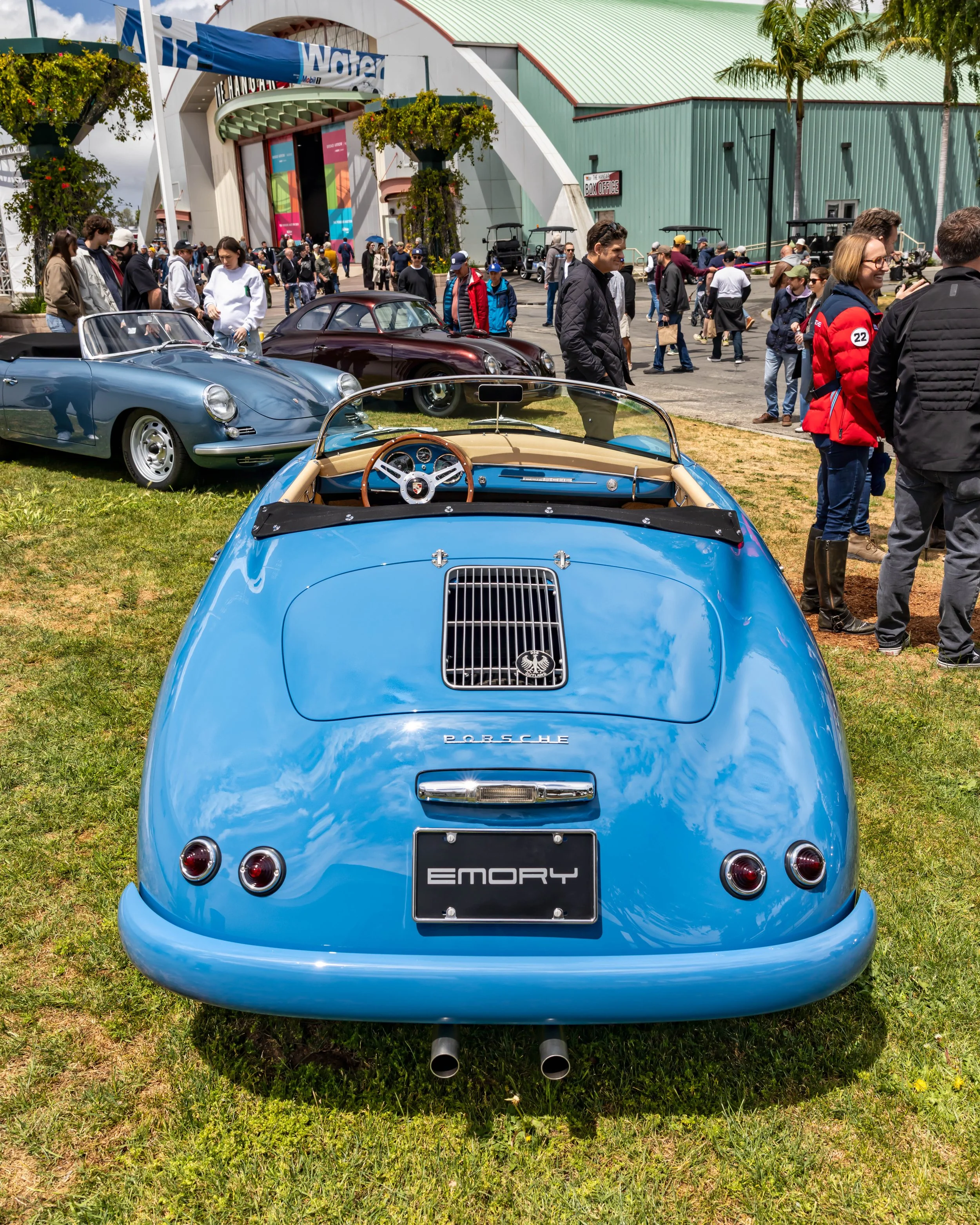 Air | Water 2025 — Emory Motorsports — OC Fairgrounds — A high-angle rear view of a light blue Emory Outlaw Porsche 356 Speedster parked on a grassy field at a car show. The car features a tan leather interior, a wood-rimmed steering wheel, and a cus