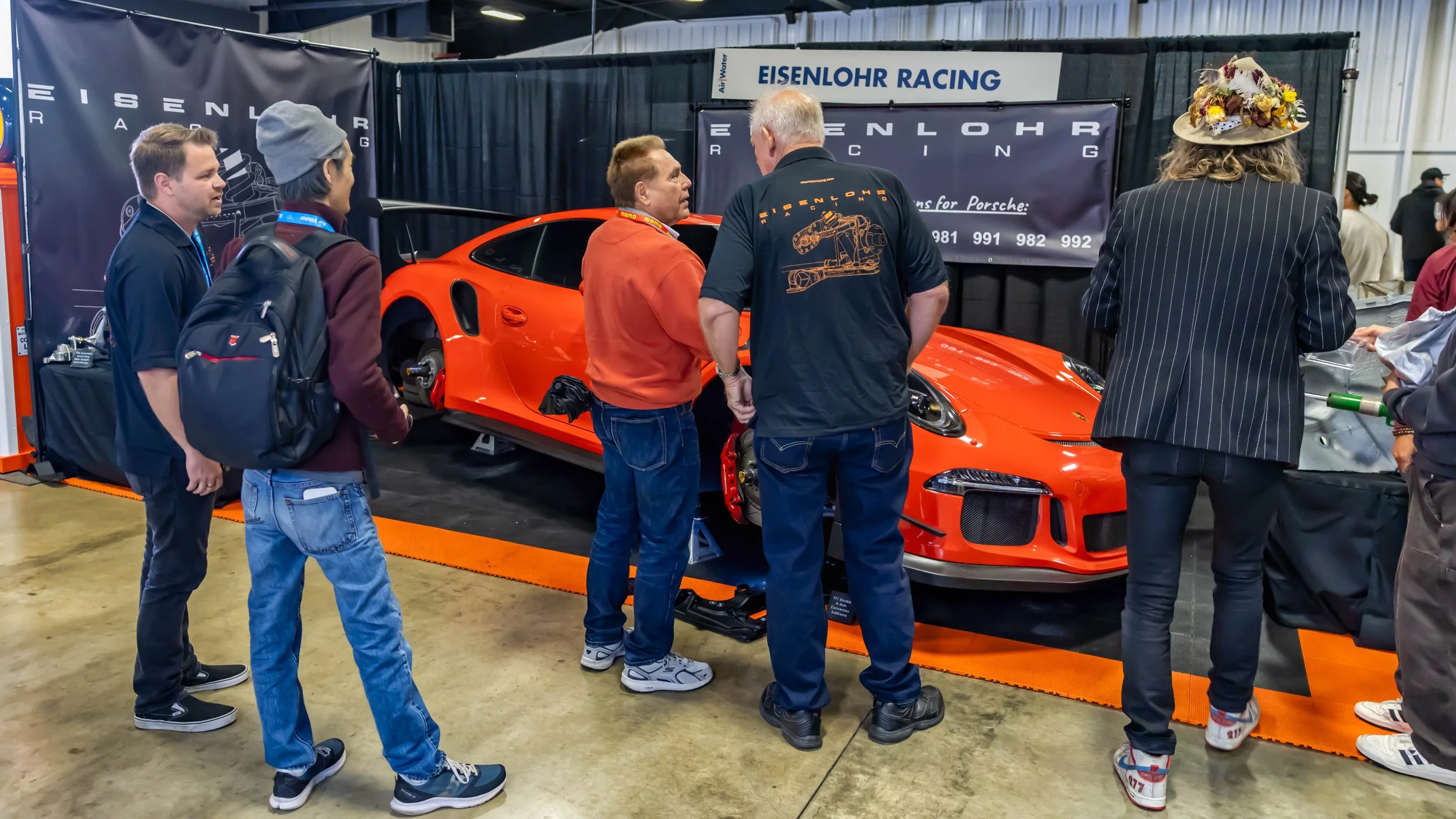 Air | Water 2025 — Orange County Fairgrounds — An orange Porsche 911 GT3 RS is displayed on jack stands with its wheels removed at the Eisenlohr Racing booth during an indoor automotive event. Several people are gathered around the car, looking at it