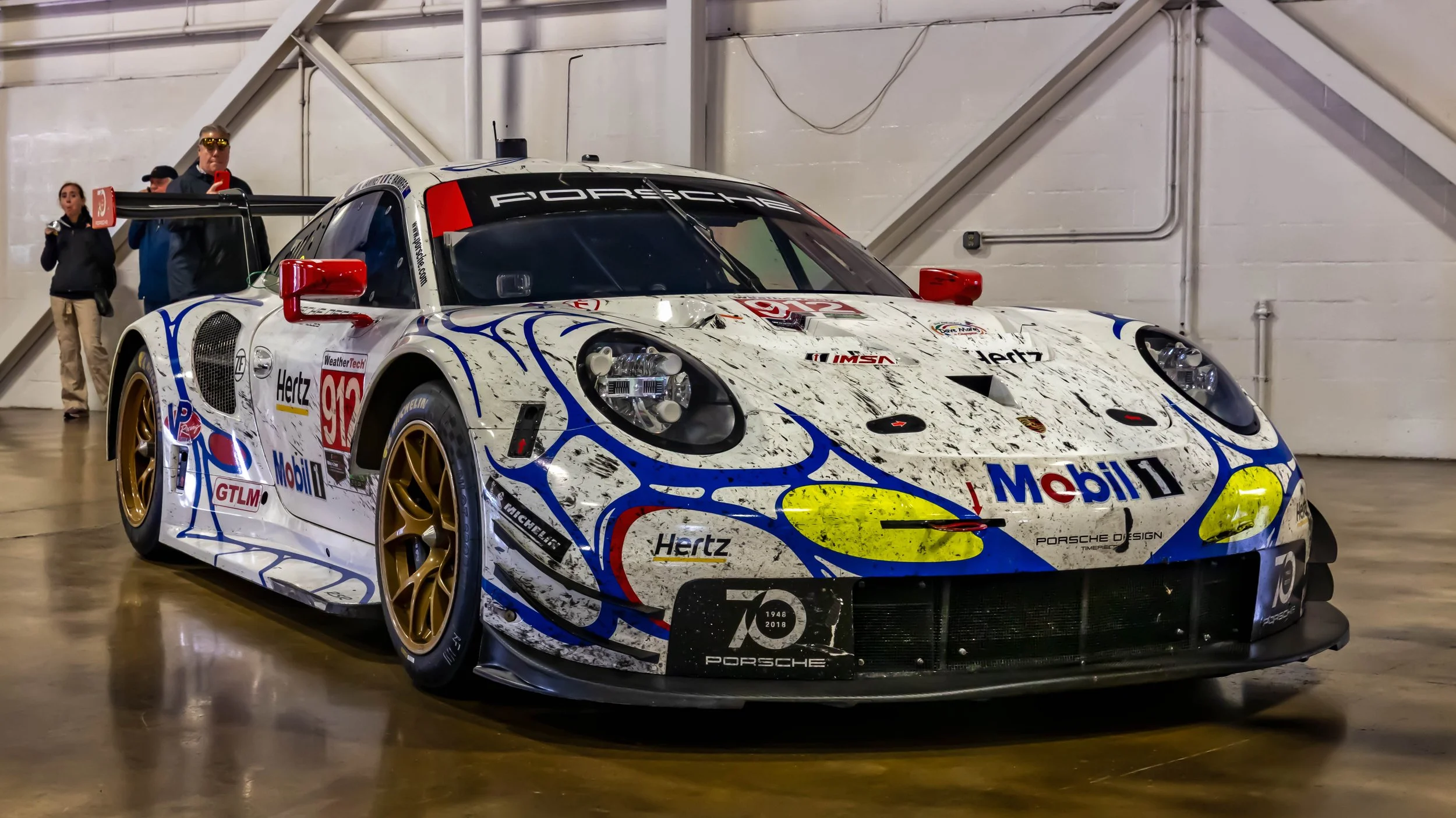 Air | Water 2025 — Orange County Fairgrounds — A front three-quarters view of the #912 Porsche 911 RSR race car, parked indoors in an exhibition hall. The car features a distinctive white, blue, red, and yellow livery sponsored by Mobil 1 and Hertz, 