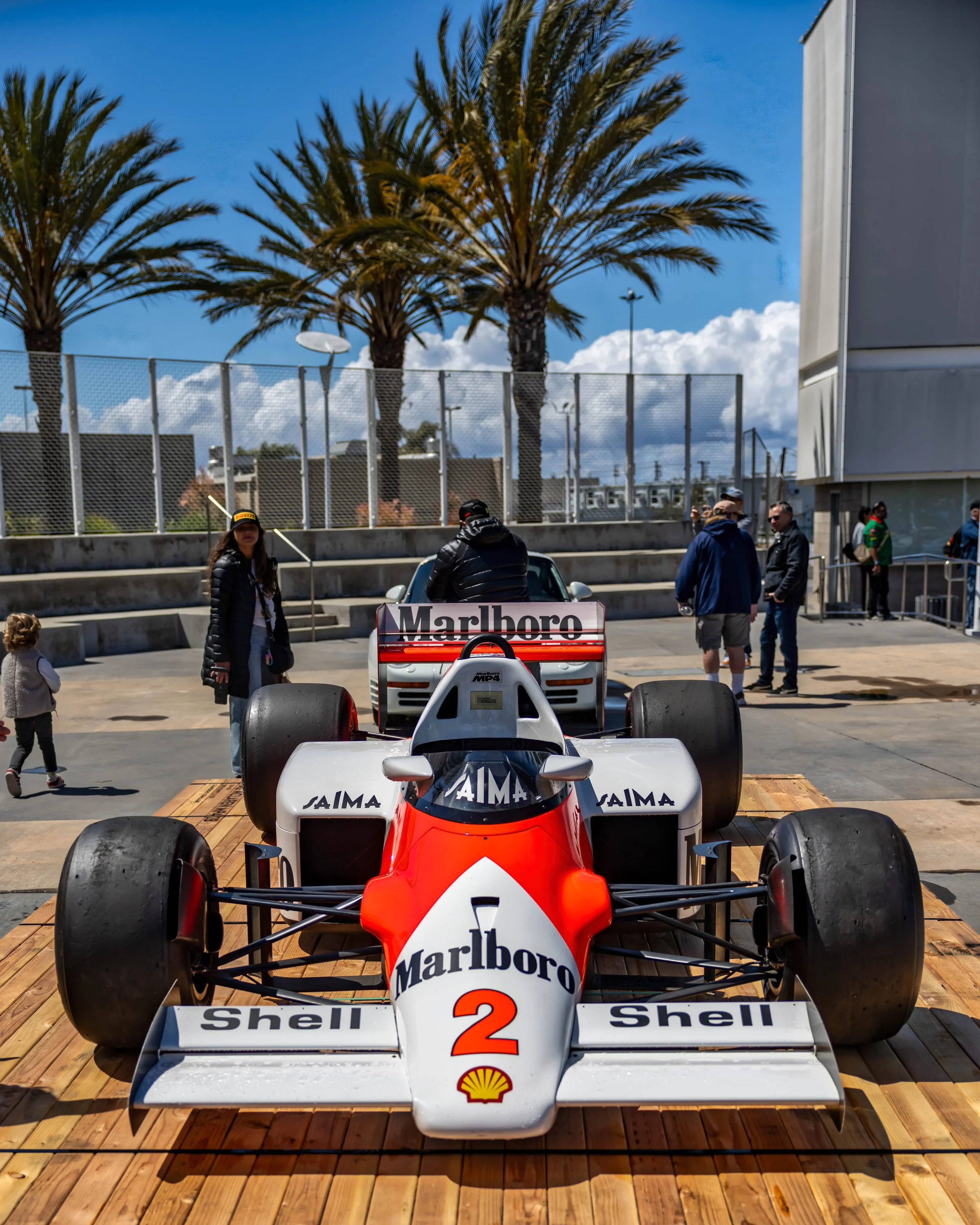 Air | Water 2025 — Orange County Fairgrounds — A head-on, low-angle photograph of a McLaren MP4/2 Formula 1 car, number 2, in its iconic red and white Marlboro livery. The race car is displayed on a wooden platform in an outdoor plaza, with palm tree
