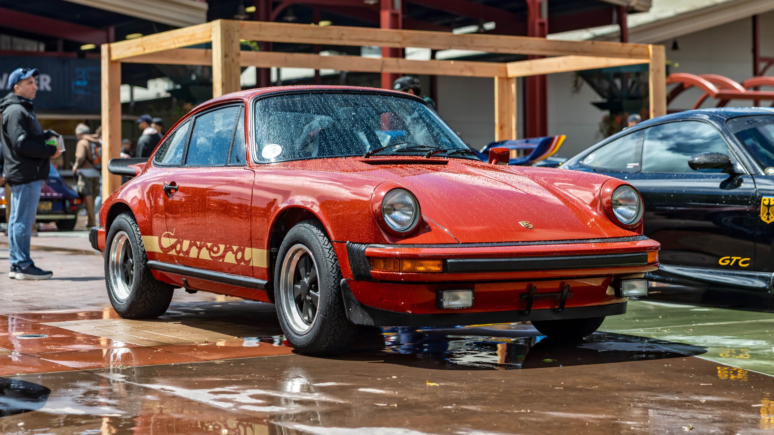 Air | Water 2025 — Orange County Fairgrounds — A metallic red Porsche 911 Carrera from the mid-1970s is parked on a wet, tiled surface at an outdoor car event. The car, covered in water droplets, features gold 'Carrera' side decals, a prominent 'whal
