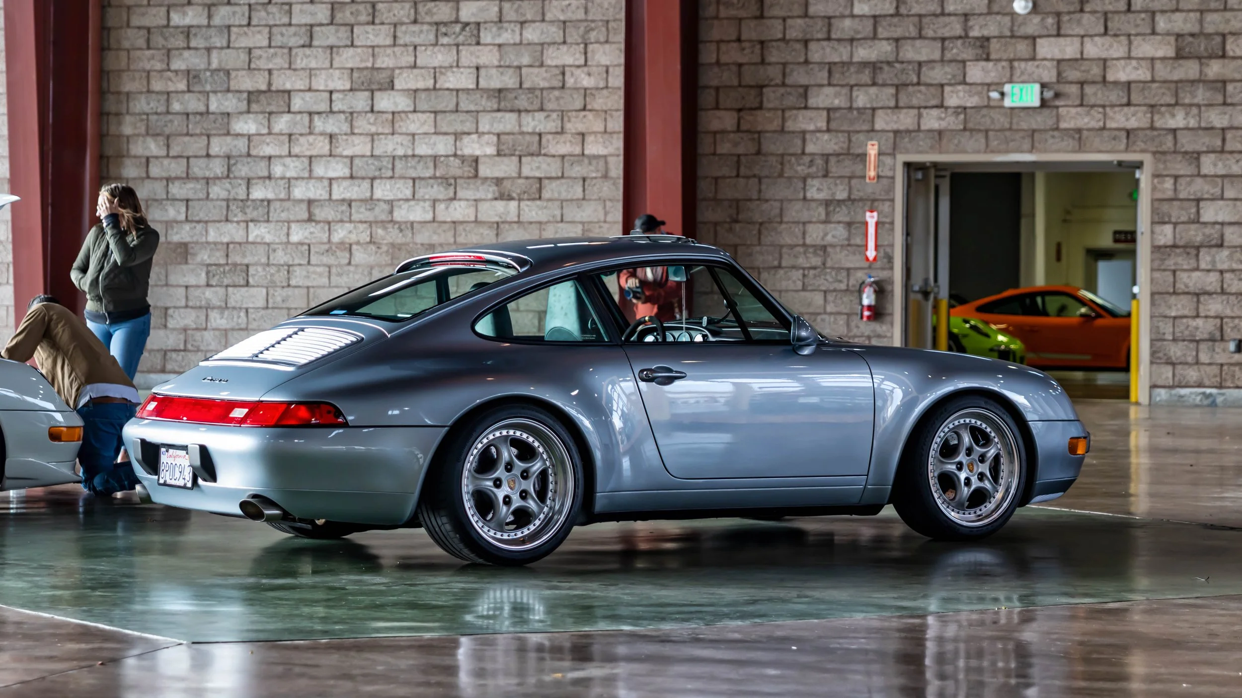 Air | Water 2025 — Orange County Fairgrounds — A silver metallic Porsche 911 Carrera of the 993 generation is parked indoors on a reflective polished concrete floor. The car is captured from a rear three-quarter angle, showcasing its coupe silhouette